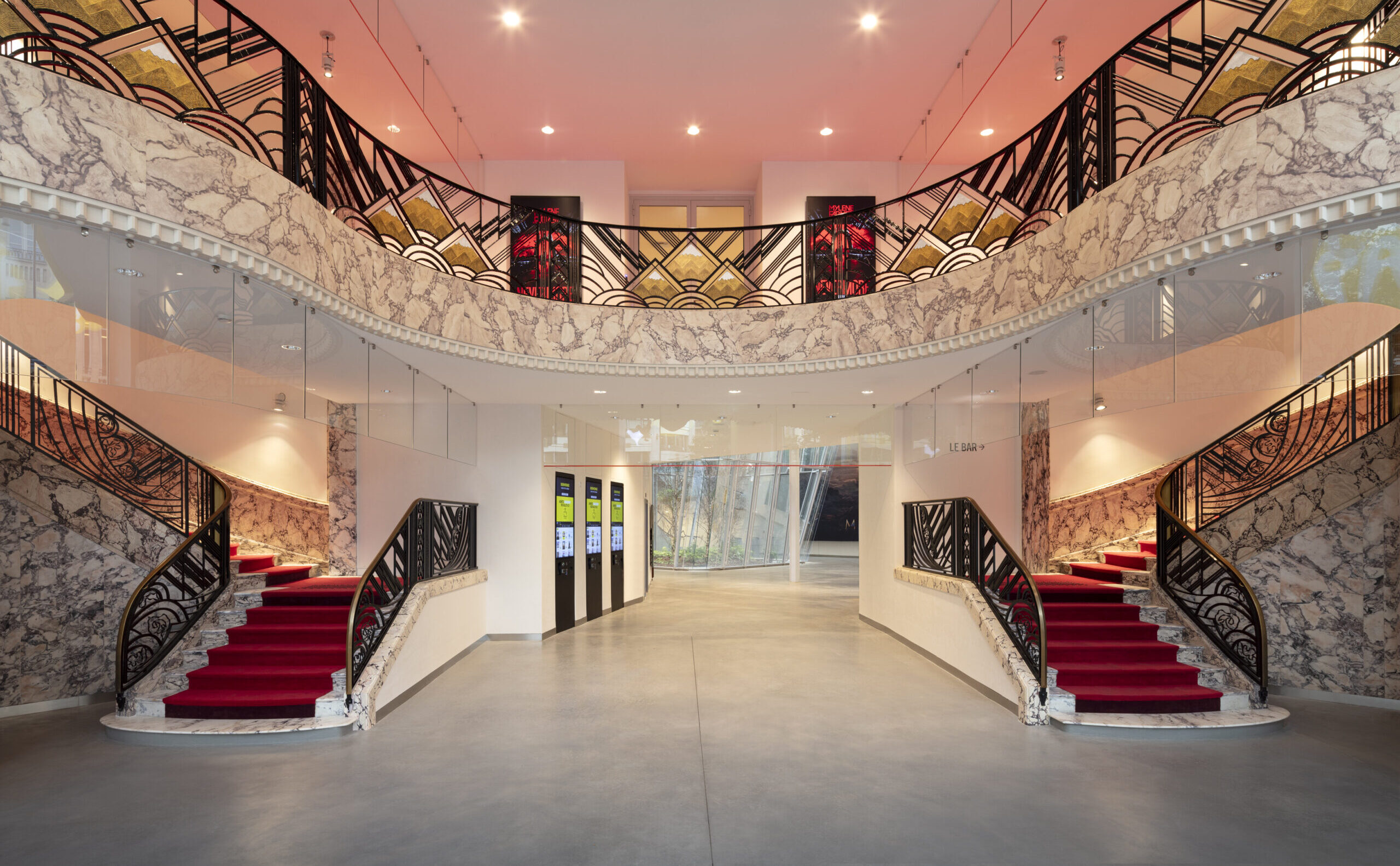 Entrée avec escalier central recouvert de tapis rouge, balustrades en métal noir décoratif et murs en marbre. Éclairage chaleureux accentuant le style art déco de l’espace.