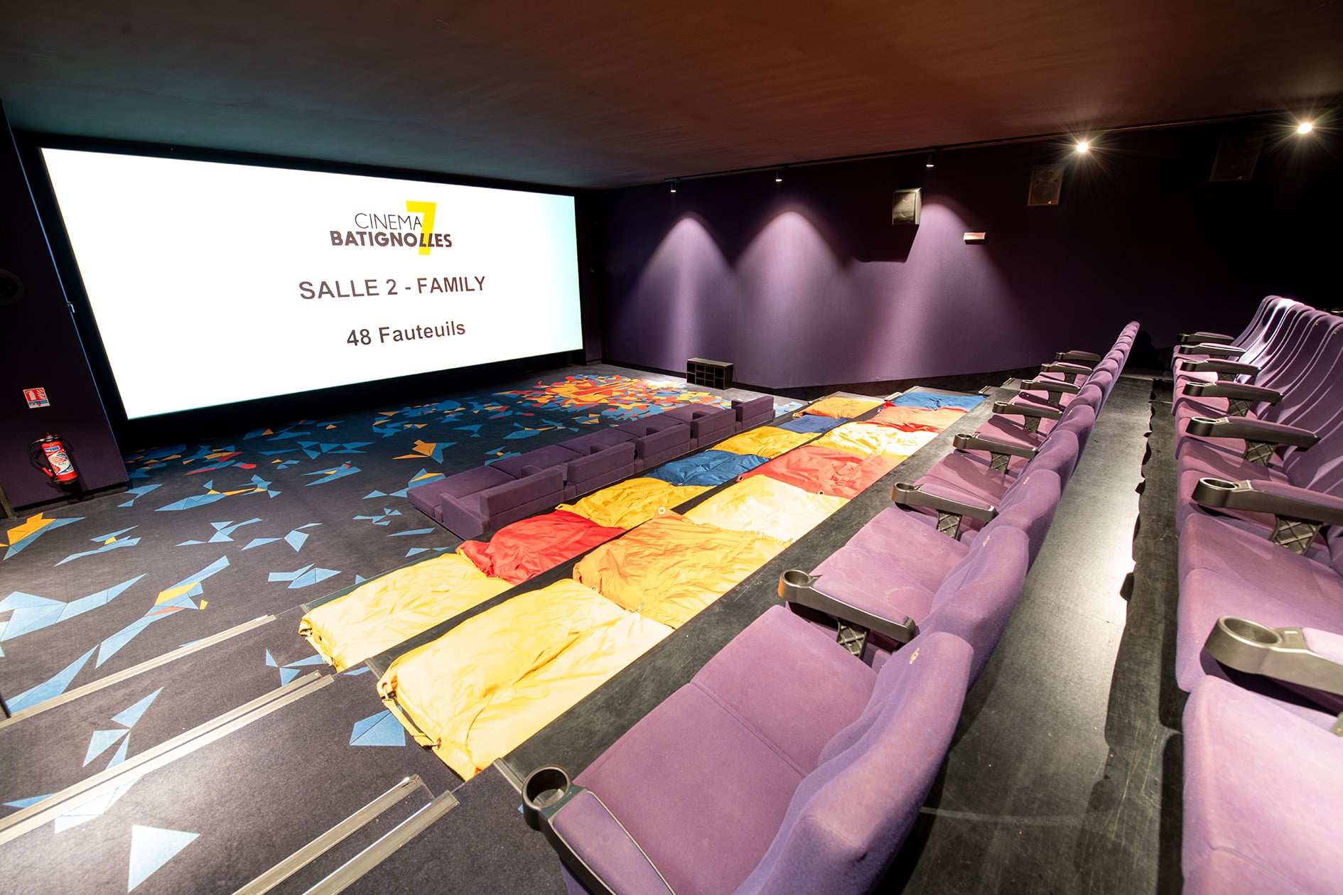 Salle familiale avec 48 fauteuils violets, matelas colorés au sol, moquette à motifs géométriques et écran large.