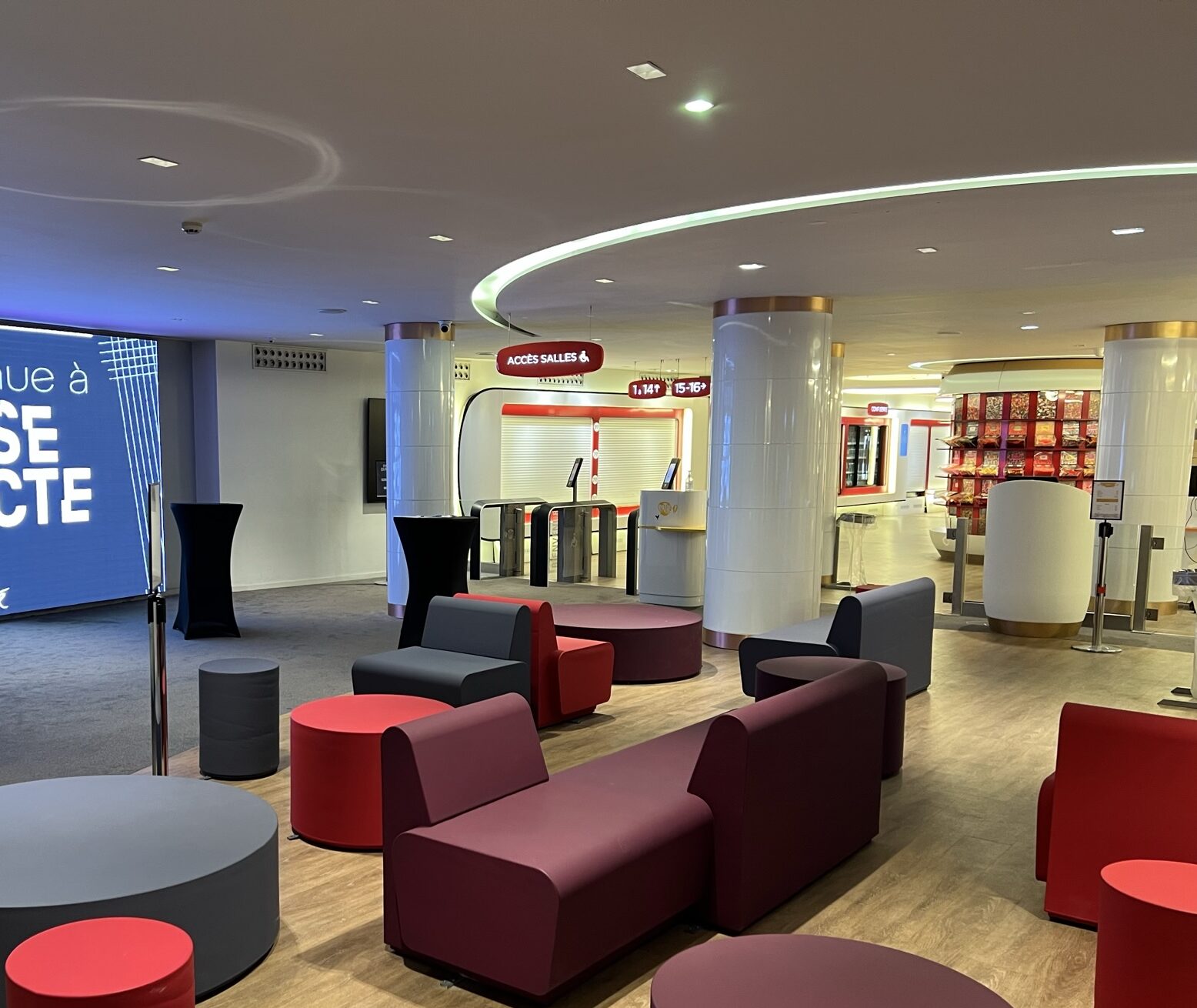 Espace de réception avec mobilier coloré composé de fauteuils et tables basses rouges, bordeaux et gris. À l’arrière, des piliers blancs, un espace avec des caisses automatiques, et un accès signalé vers les salles de cinéma. Le sol est en bois clair, et l’éclairage donne une ambiance lumineuse.