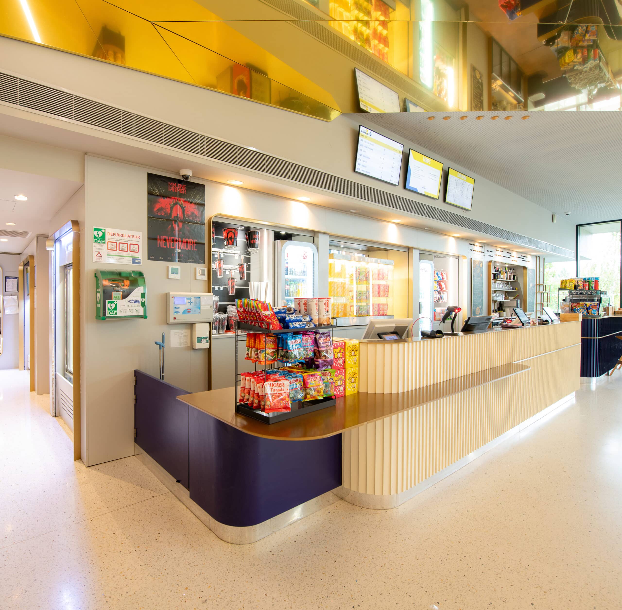 Comptoir de confiserie dans le cinéma Pathé, avec présentoir rempli de bonbons, écrans affichant les tarifs, vitrines réfrigérées et espace lumineux au design moderne.
