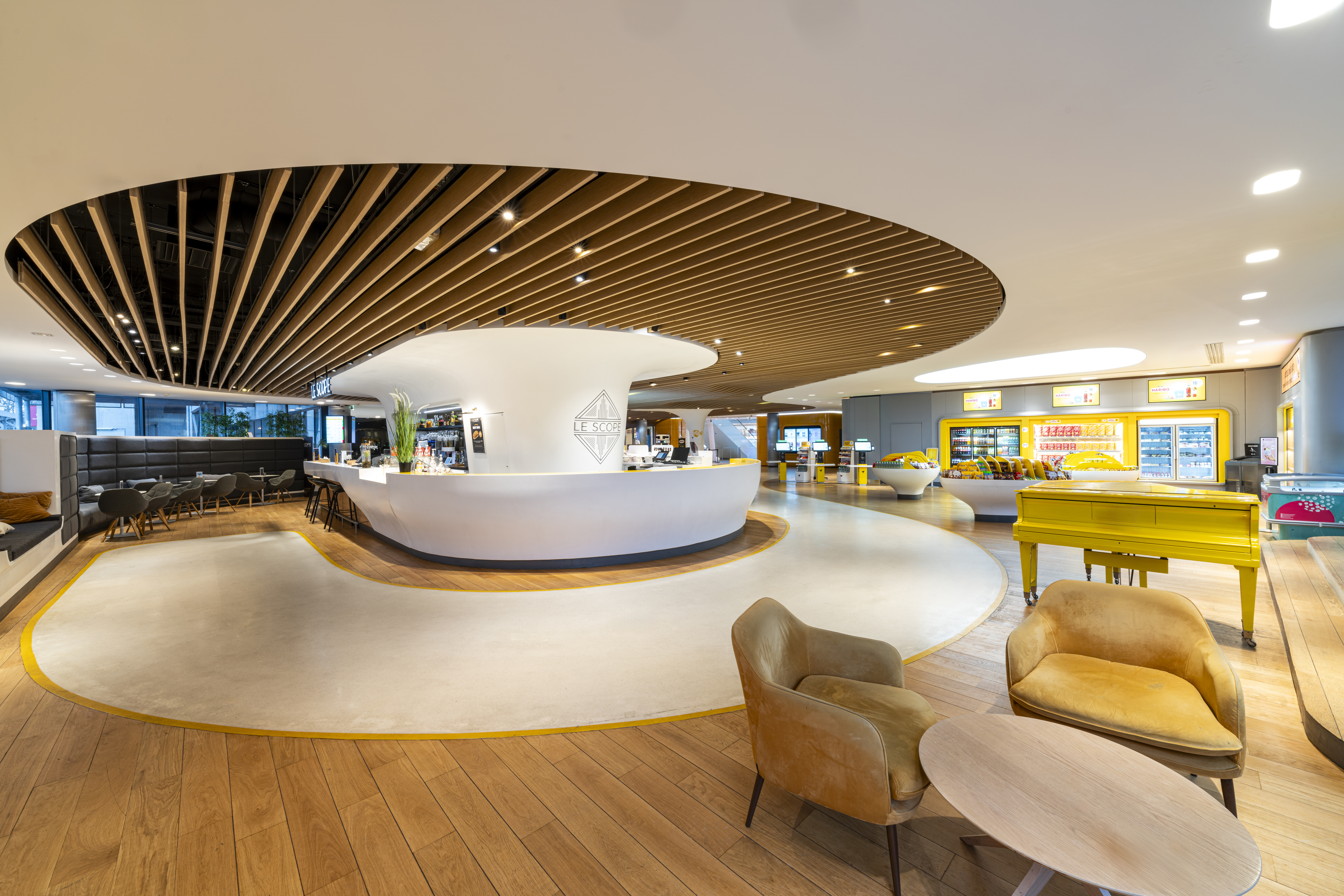 Espace clair et moderne avec comptoir arrondi blanc, plafond en bois lamellé, assises variées, piano jaune, et zone de snacks réfrigérés à l'arrière.