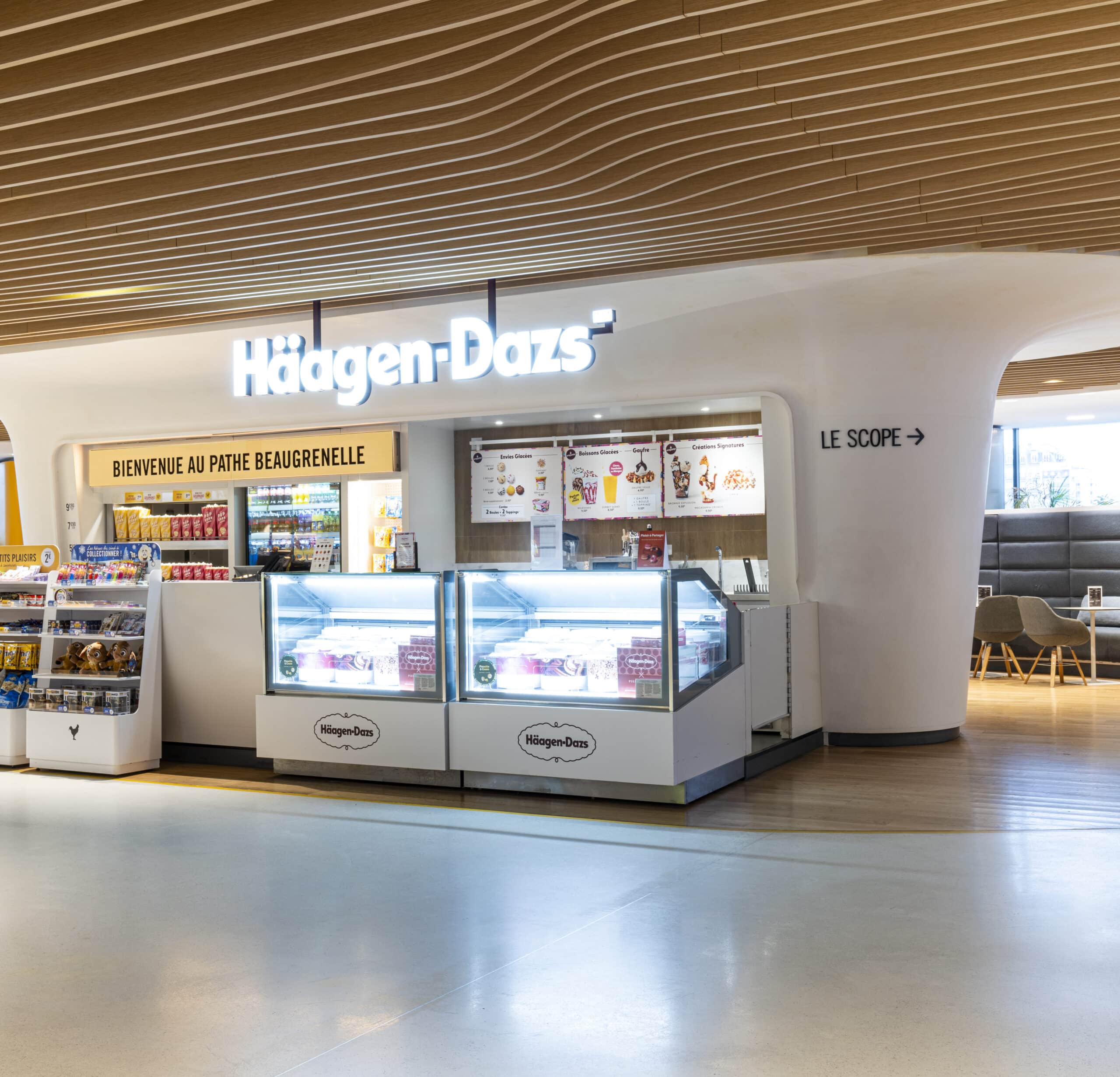 Comptoir Häagen-Dazs dans le cinéma Pathé Beaugrenelle, avec vitrines réfrigérées remplies de glaces, enseigne lumineuse et présentoirs de snacks. Décoration moderne avec plafond ondulé en bois et espace détente visible en arrière-plan.