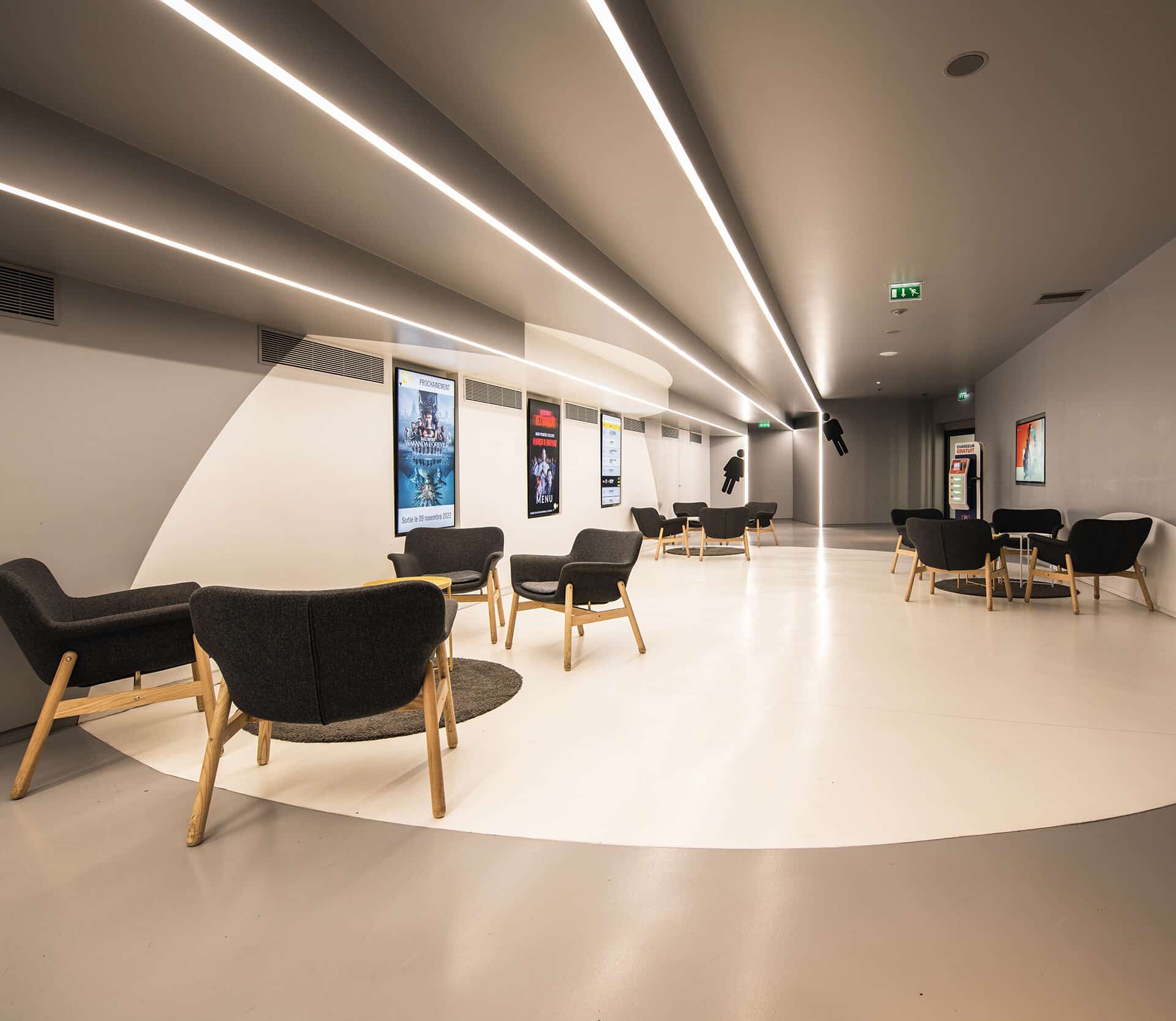 Espace de réception moderne avec sol clair et mobilier composé de fauteuils noirs à structure en bois. Des affiches de films sont accrochées au mur, sous un éclairage lumineux intégré au plafond. L’ambiance est sobre et épurée.