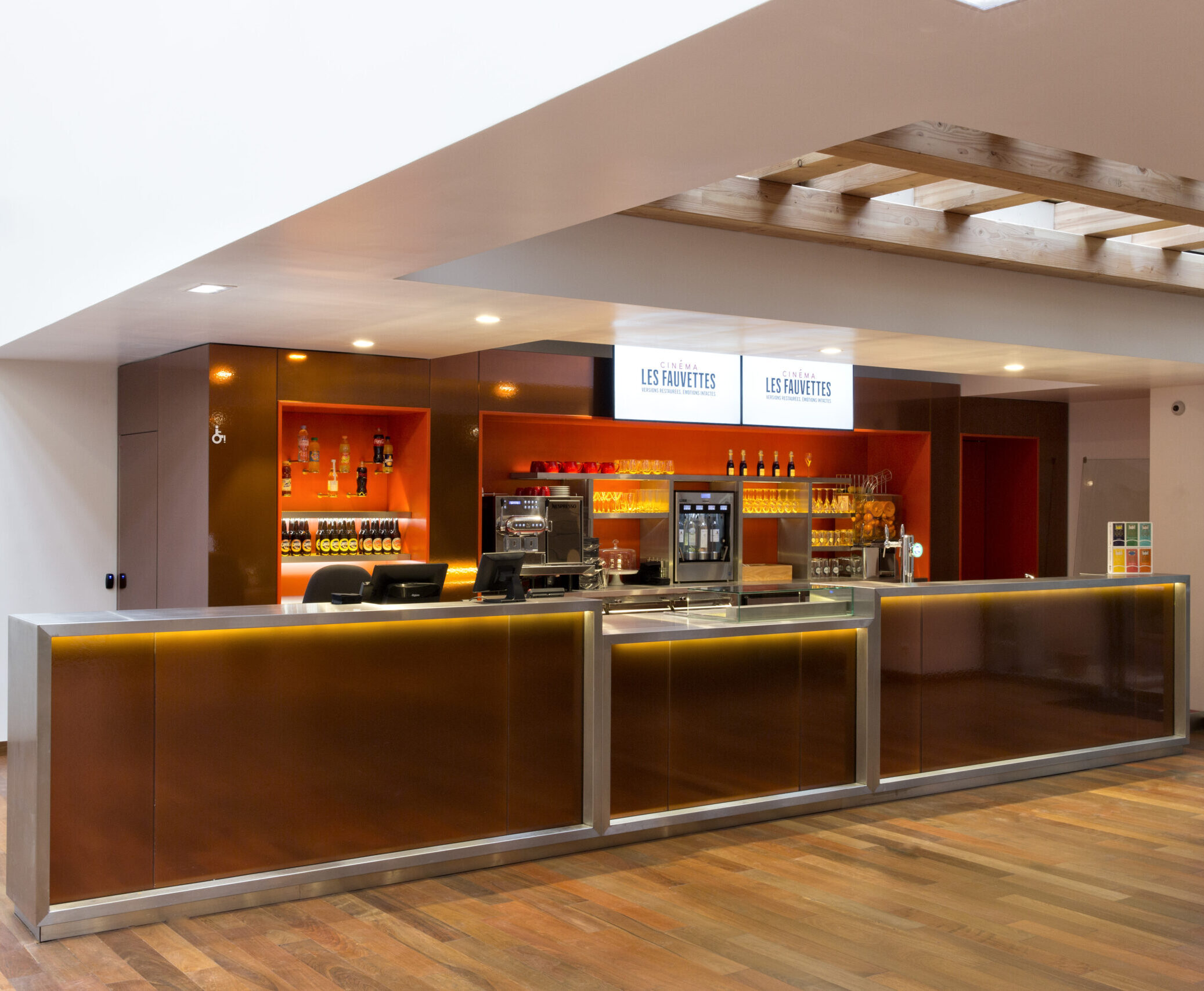 Comptoir de service moderne dans un espace lumineux, avec un sol en parquet bois foncé. À l'arrière, des étagères éclairées exposent des bouteilles, des verres et une machine à café. Au-dessus du comptoir, deux écrans affichent le logo 
