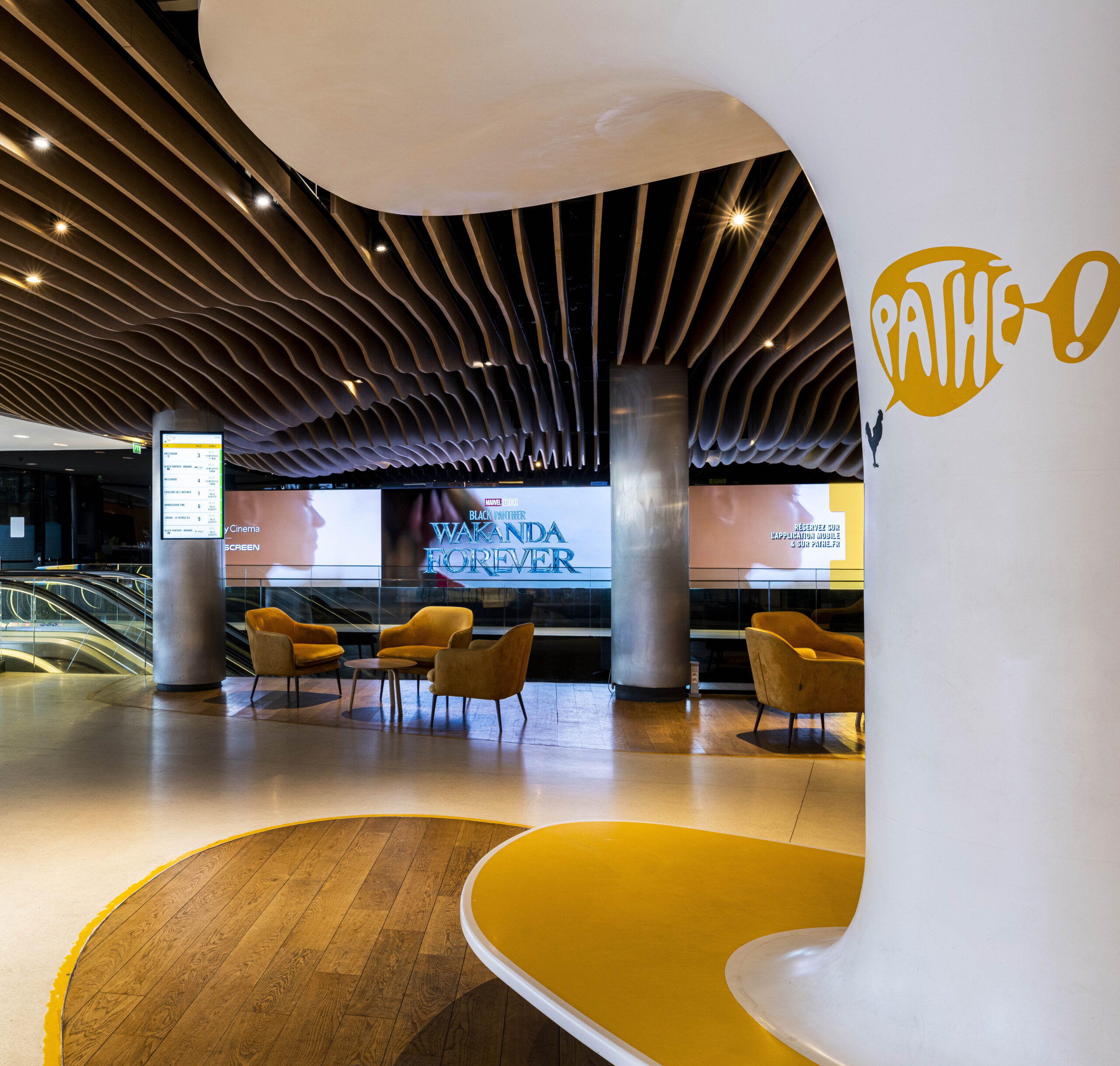 Espace cinématographique Pathé avec fauteuils jaunes autour de petites tables, sol combinant parquet et béton ciré, et plafond ondulé moderne. Affiche lumineuse du film 