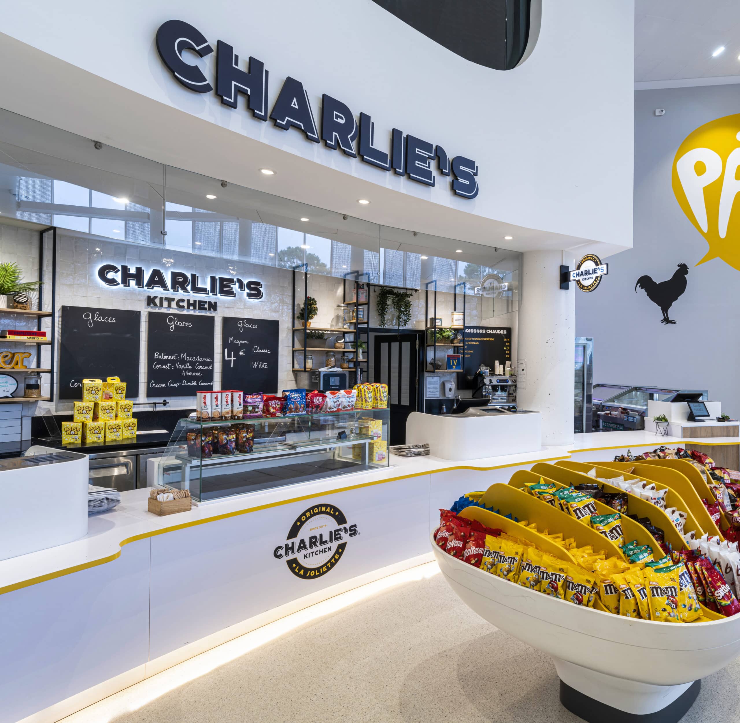 Comptoir Charlie's Kitchen avec enseigne lumineuse, étagères garnies de snacks, vitrine de glaces et présentoir de bonbons en forme de bol. Décor moderne avec murs blancs, plantes et grandes fenêtres laissant passer la lumière naturelle. Logo Pathé visible sur le mur.