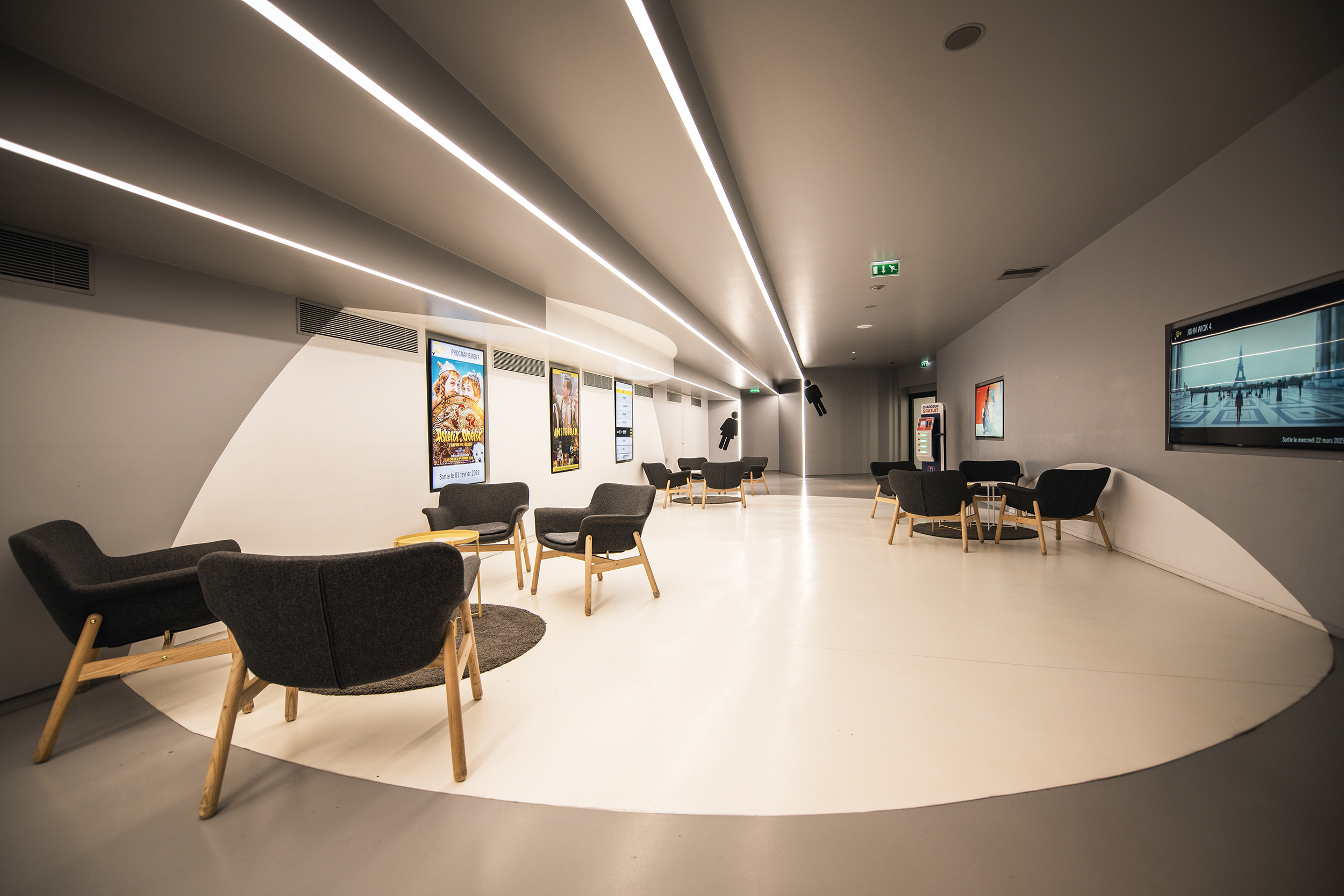 Salon lumineux avec sol blanc, éclairage en lignes au plafond, fauteuils modernes en tissu, tables basses et affiches de films sur les murs.