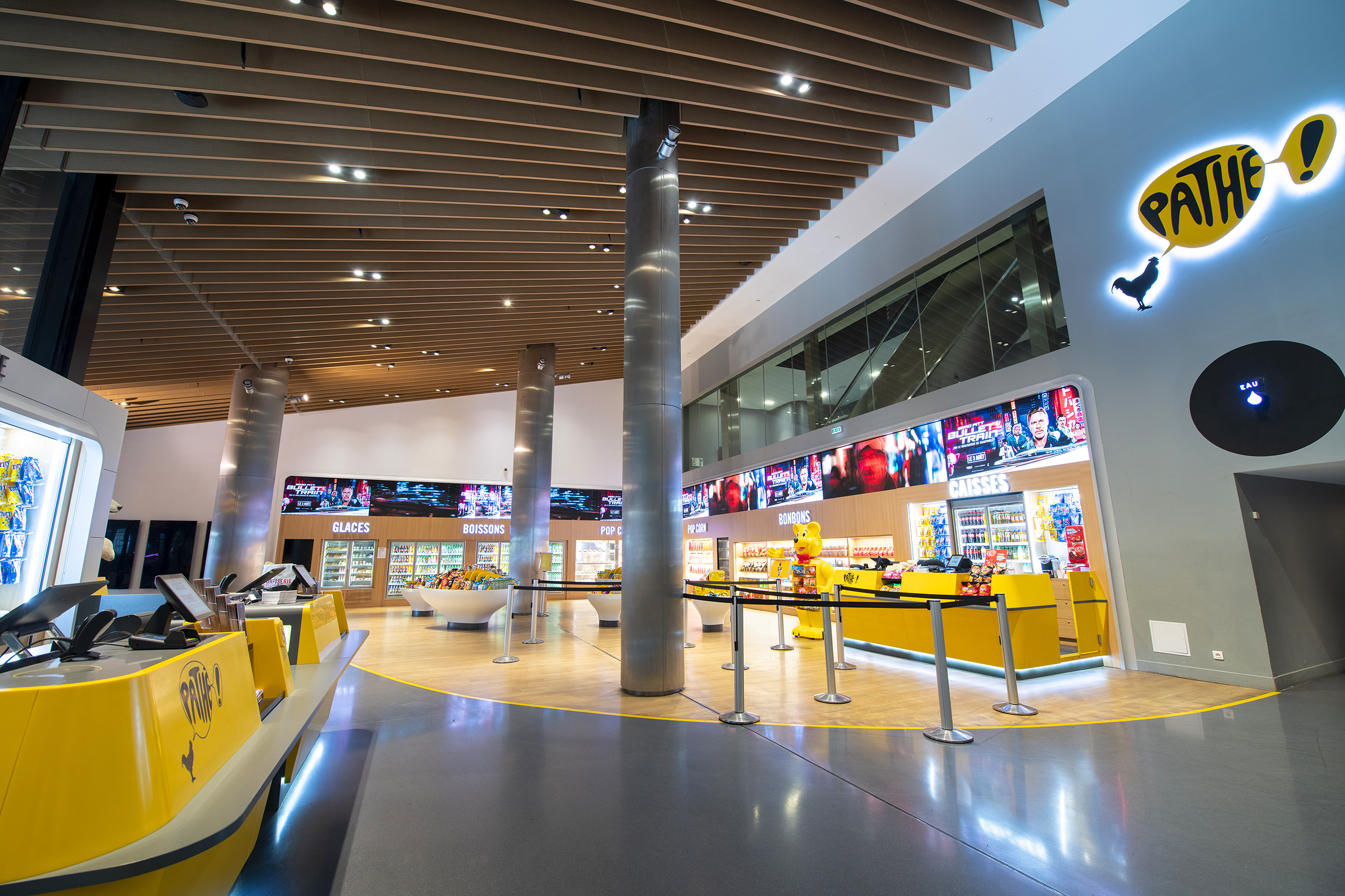 Hall de cinéma spacieux avec comptoirs lumineux jaunes, panneaux d’affichage, vitrines réfrigérées pour snacks et boissons, et plafond en bois lamellé.