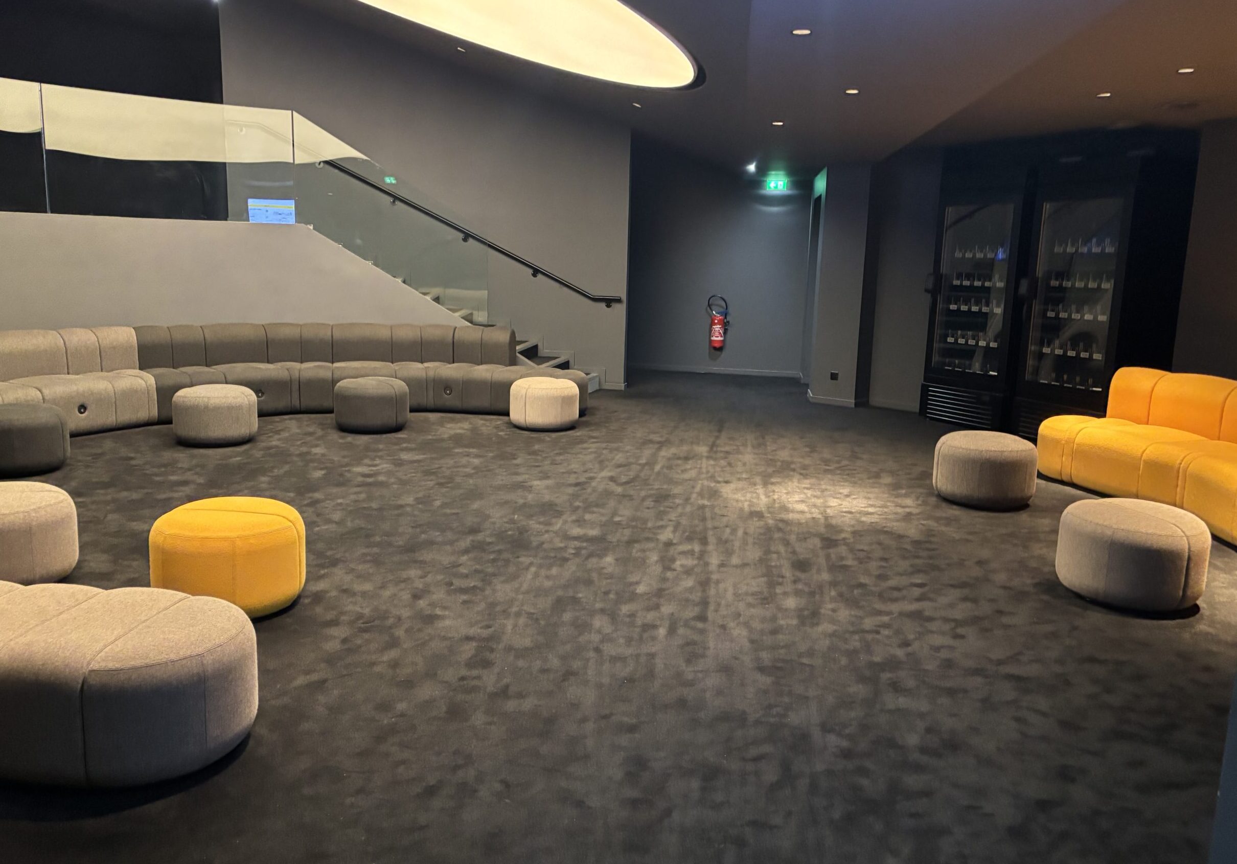 Espace lounge avec moquette grise, sièges arrondis gris et jaunes, canapé en arc de cercle, et escalier en verre menant à l'étage
