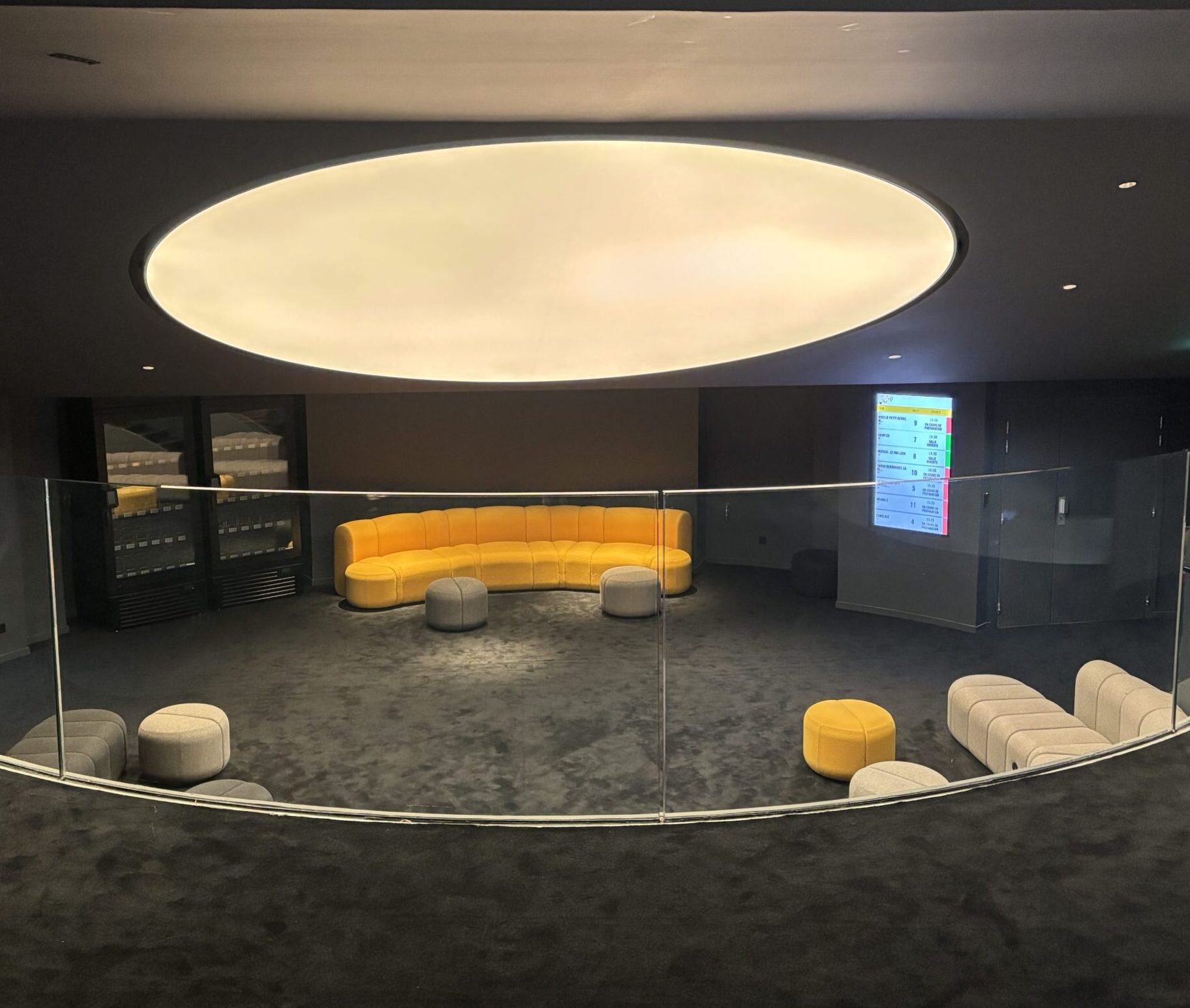 Espace de réception avec canapé arrondi jaune vif, poufs gris et moquette sombre. Luminaire circulaire créant une ambiance douce et moderne. Écran mural affichant des informations sur les salles. Style épuré et chaleureux.