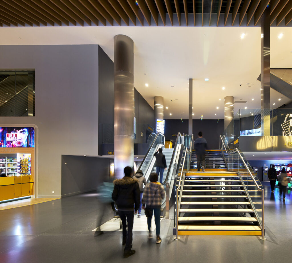 Hall animé avec escalators et escalier central, colonnes métalliques, plafond lamellé, et zones de caisses et snacks en arrière-plan.