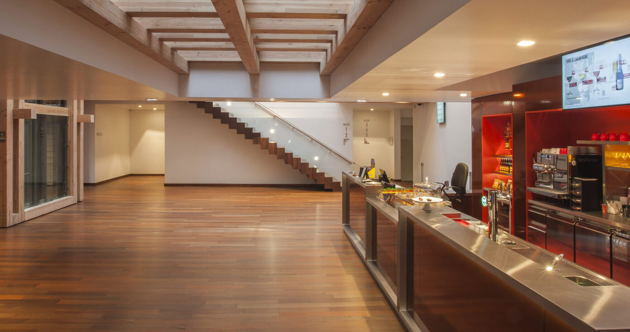 Espace moderne avec parquet en bois, un bar équipé d'une machine à café et de boissons. Escalier design avec rampe en verre, éclairage chaleureux et structure en bois visible au plafond.