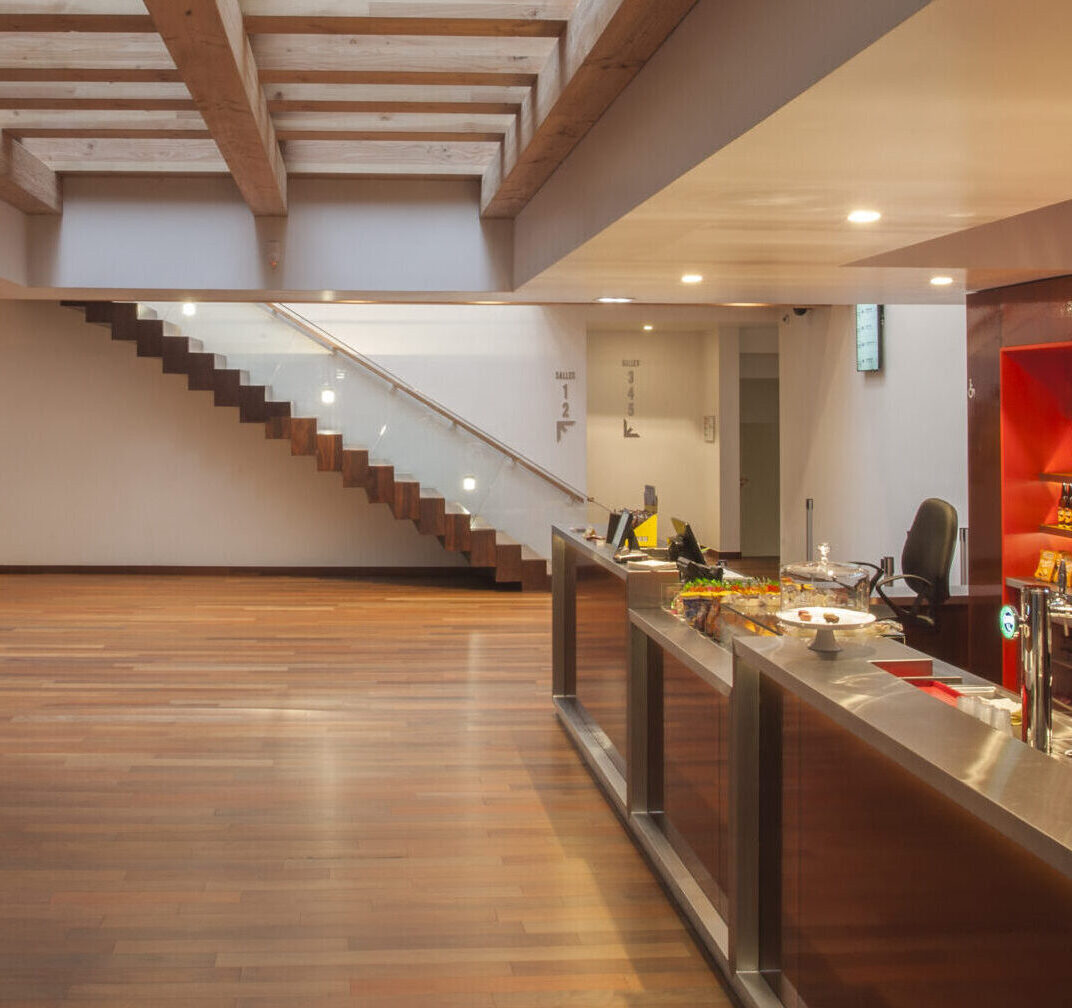 Espace moderne avec parquet en bois, un bar équipé d'une machine à café et de boissons. Escalier design avec rampe en verre, éclairage chaleureux et structure en bois visible au plafond.
