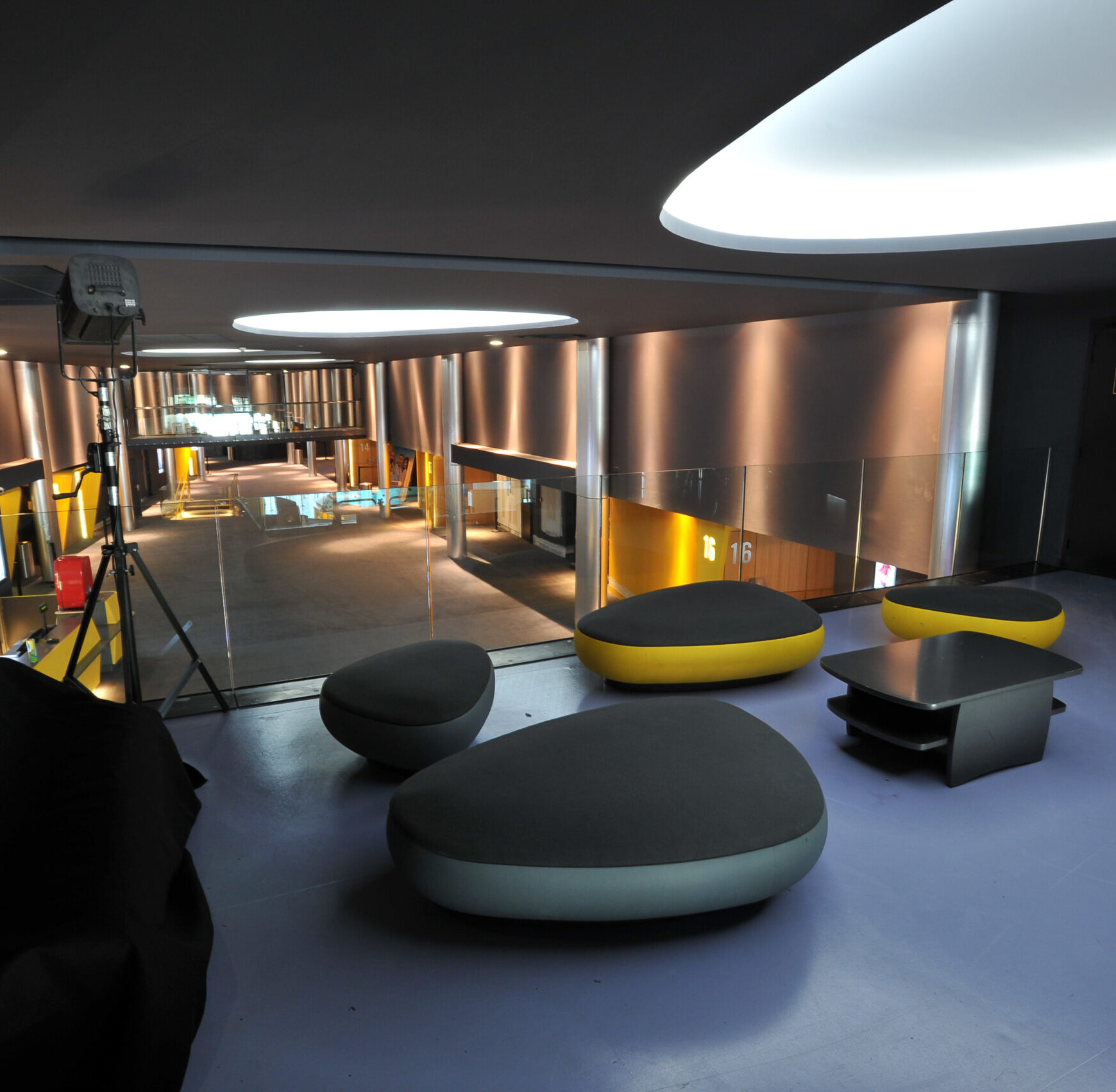 Espace intérieur avec un mobilier moderne composé de sièges arrondis noirs et jaunes, ainsi que d’une table basse foncée. Le sol est violet et le plafond est équipé de larges luminaires circulaires diffusant une lumière douce. À l’arrière, on aperçoit une mezzanine donnant sur une zone avec des murs métalliques brillants et des numéros de salles.