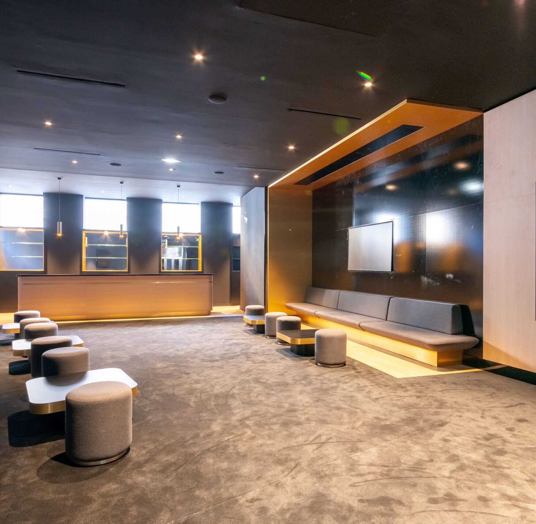 Espace de réception avec un plafond sombre éclairé par des spots. Le sol est recouvert de moquette grise, sur laquelle sont disposés des poufs gris et des tables basses à bords dorés. À gauche, une banquette grise est accolée à un mur noir brillant. À l’arrière, un ascenseur en acier inoxydable et des portes offrent des accès pratiques.