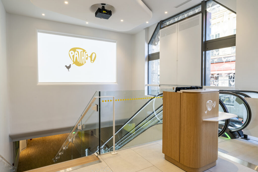 Entrée lumineuse du cinéma avec mobilier en bois, escalators et logo Pathé en projection.