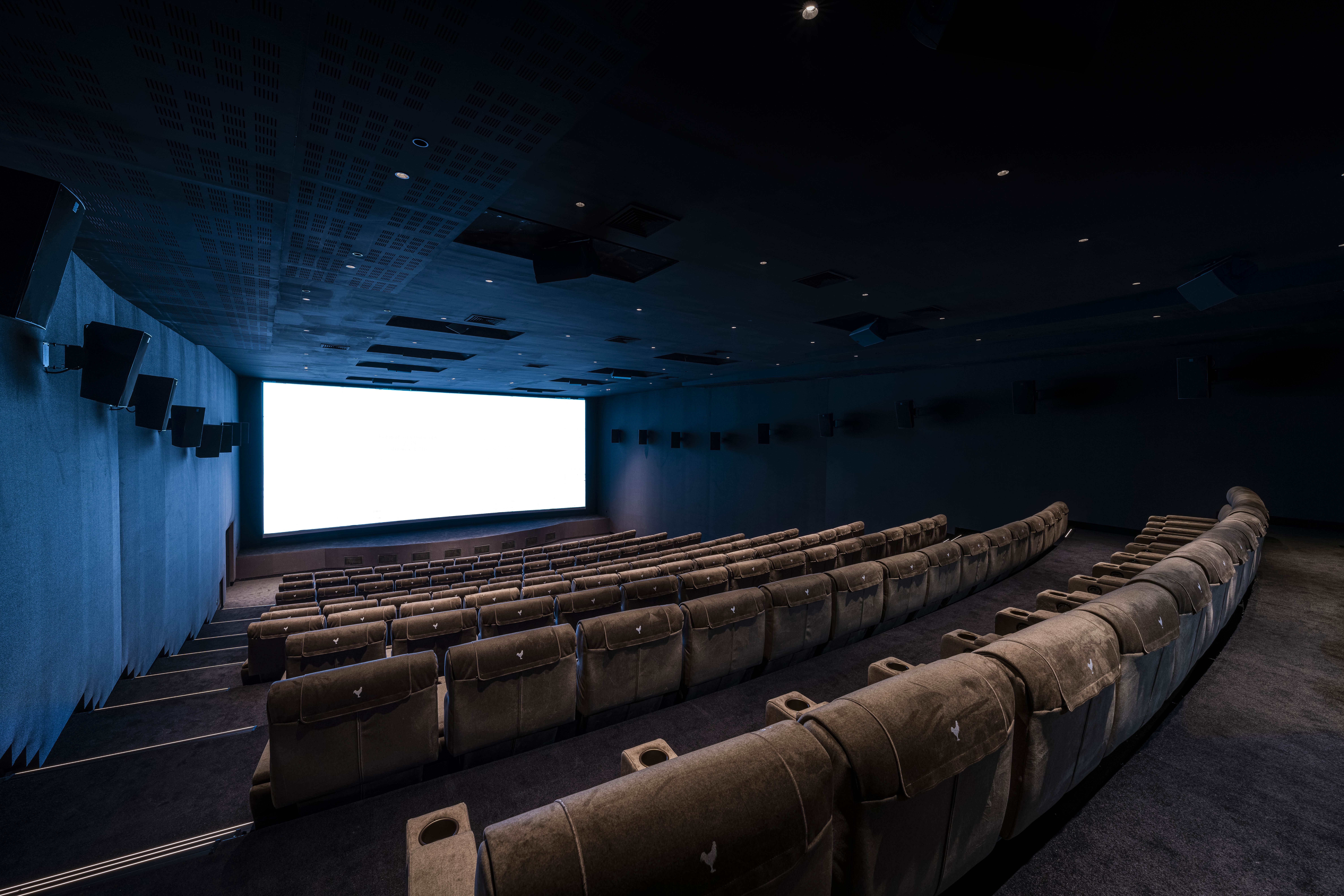 Salle de cinéma moderne avec écran blanc lumineux, rangées de fauteuils confortables en tissu beige et murs équipés de haut-parleurs.