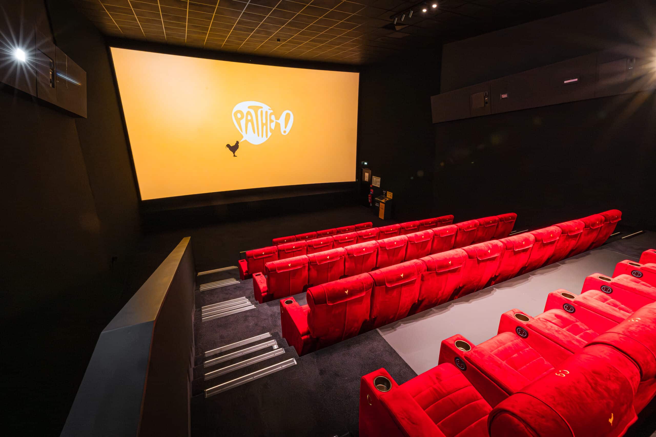 Vue depuis l’arrière d'une salle de cinéma avec ses rangées de fauteuils en velours rouges orientés vers l’écran avec le logo Pathé.