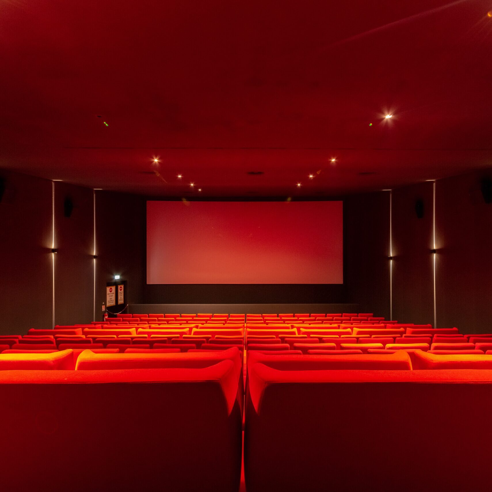 Vue depuis l’arrière d'une salle de cinéma avec ses rangées de fauteuils en velours rouges orientés vers l’écran.