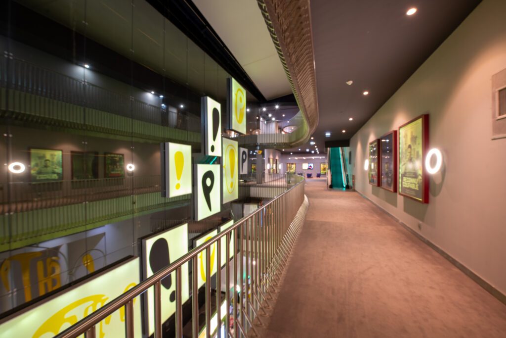 Couloir avec moquette beige, rampes métalliques, affiches lumineuses suspendues et éclairage moderne dans une ambiance élégante