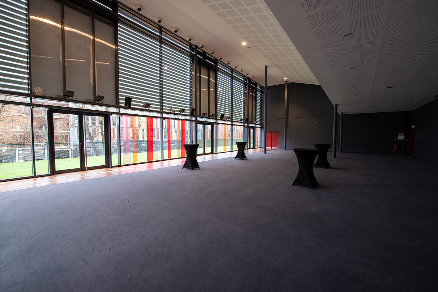 Salle de réception spacieuse avec grandes baies vitrées équipées de stores horizontaux, laissant entrevoir un extérieur coloré aux teintes rouges et jaunes. Sol en moquette sombre et petites tables hautes drapées de noir disposées dans l’espace. Ambiance épurée et lumineuse.