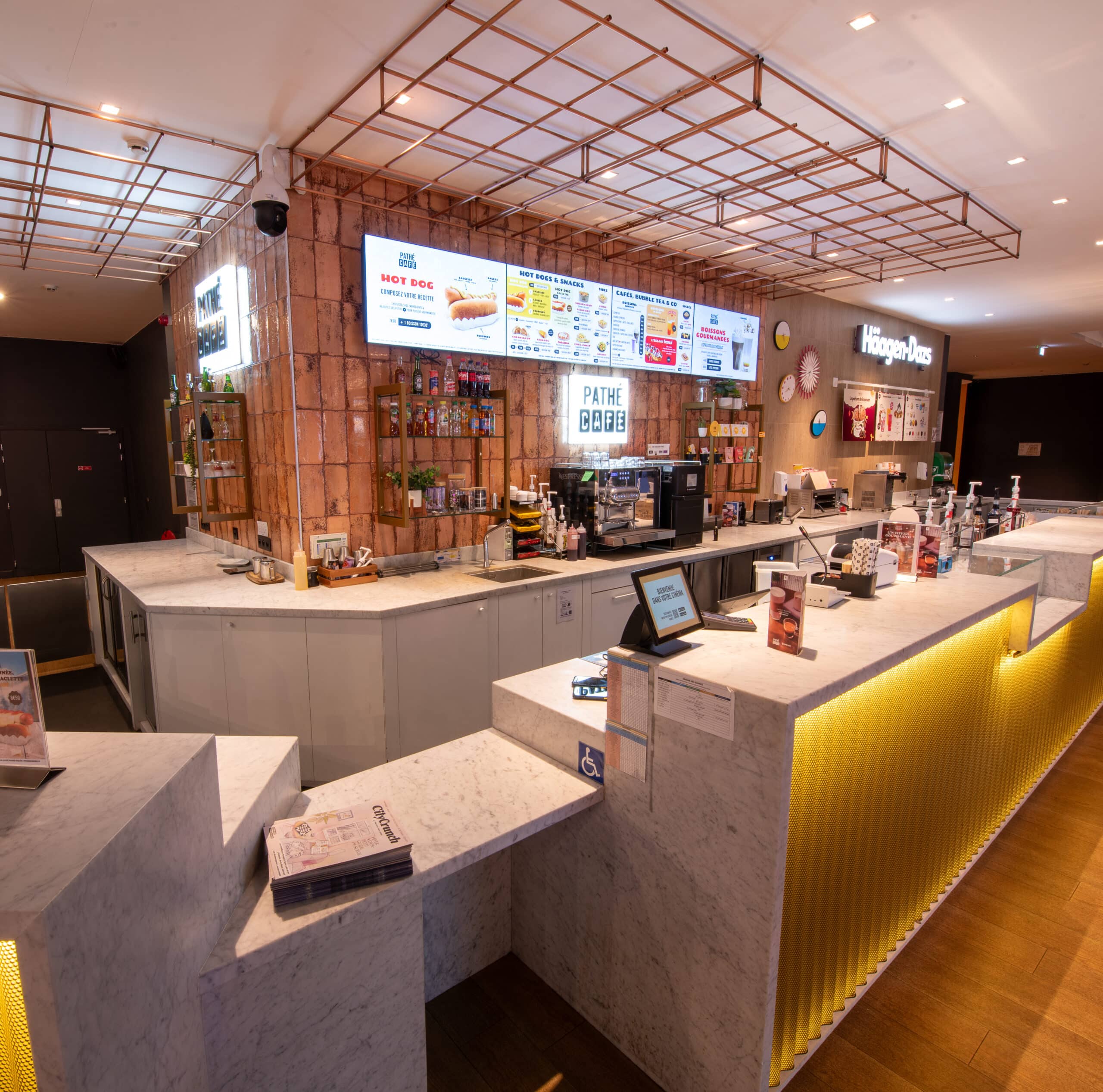 Comptoir en marbre lumineux du Pathé Café et Häagen-Dazs avec affichages numériques, étagères en briques contenant des produits, machines à café et espace accessible. Le plafond est doté d'une grille en métal doré moderne, et le sol en bois clair crée une ambiance chaleureuse.