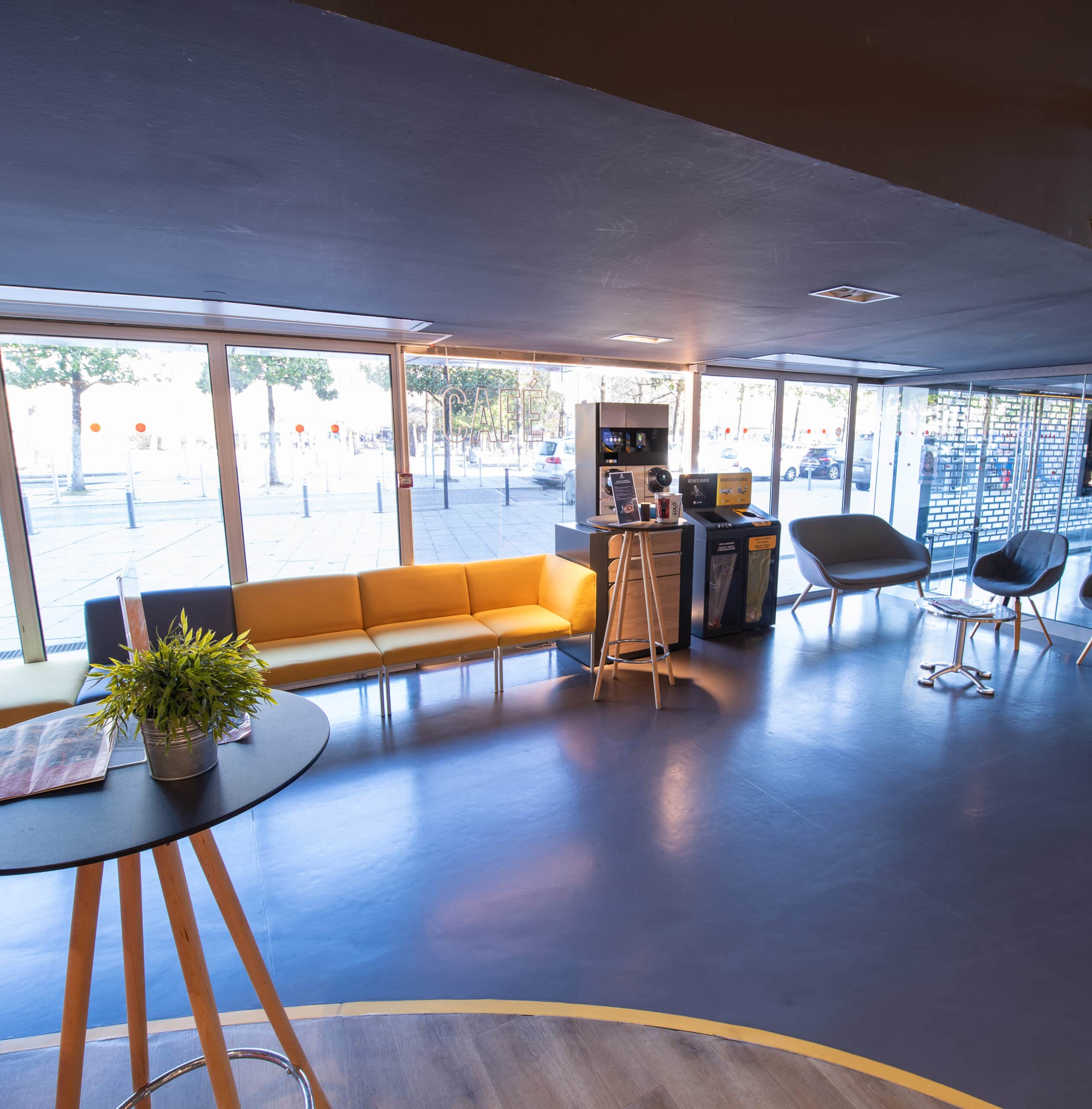 Espace de réception du cinéma à la lumière du jour grâce à ses baies vitrées, équipé d'un mobilier de réception jaune et gris.