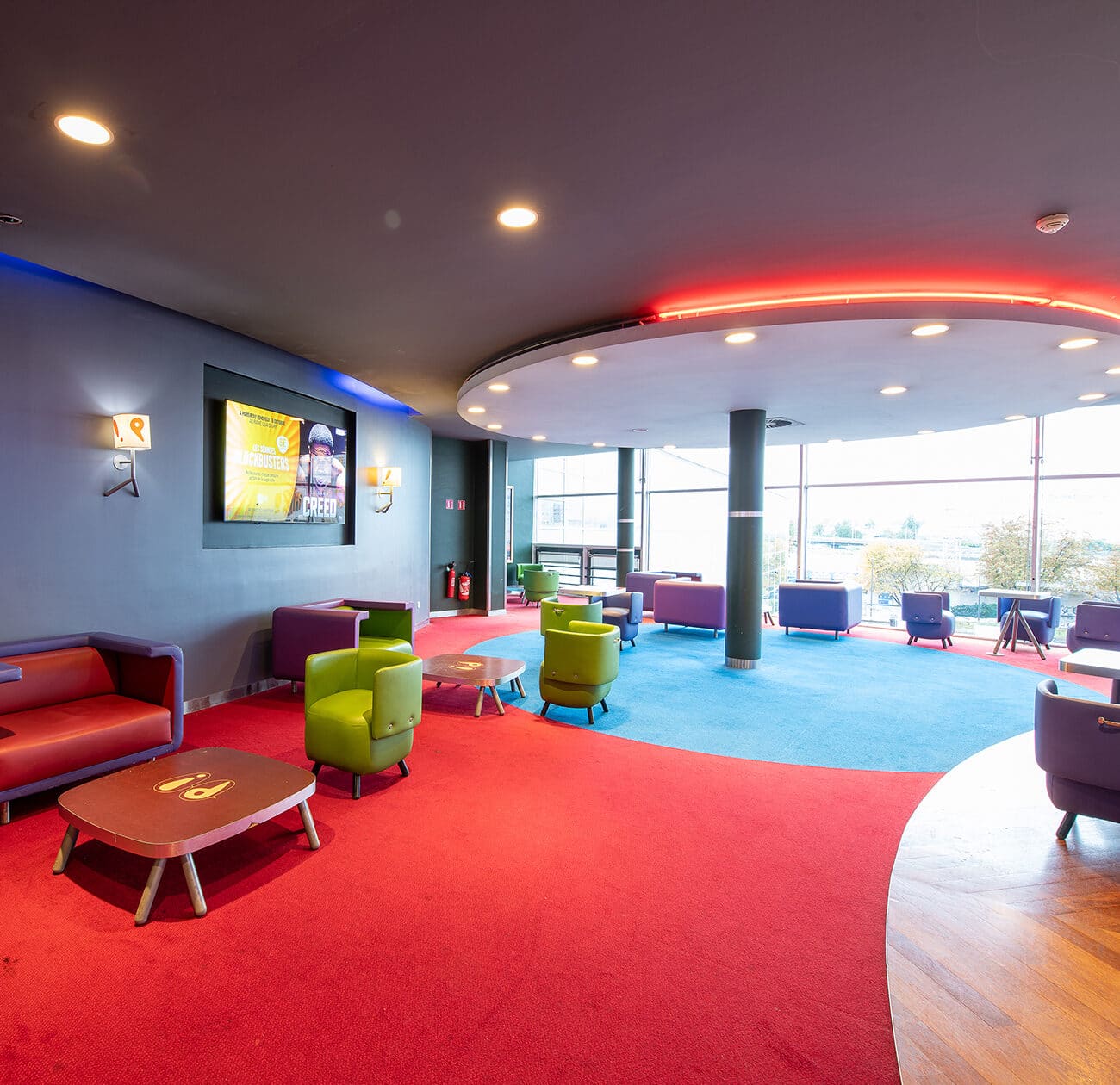 Espace cocktail du cinéma, éclairé par des lumières colorées et aménagé avec du mobilier de réception vert et violet.