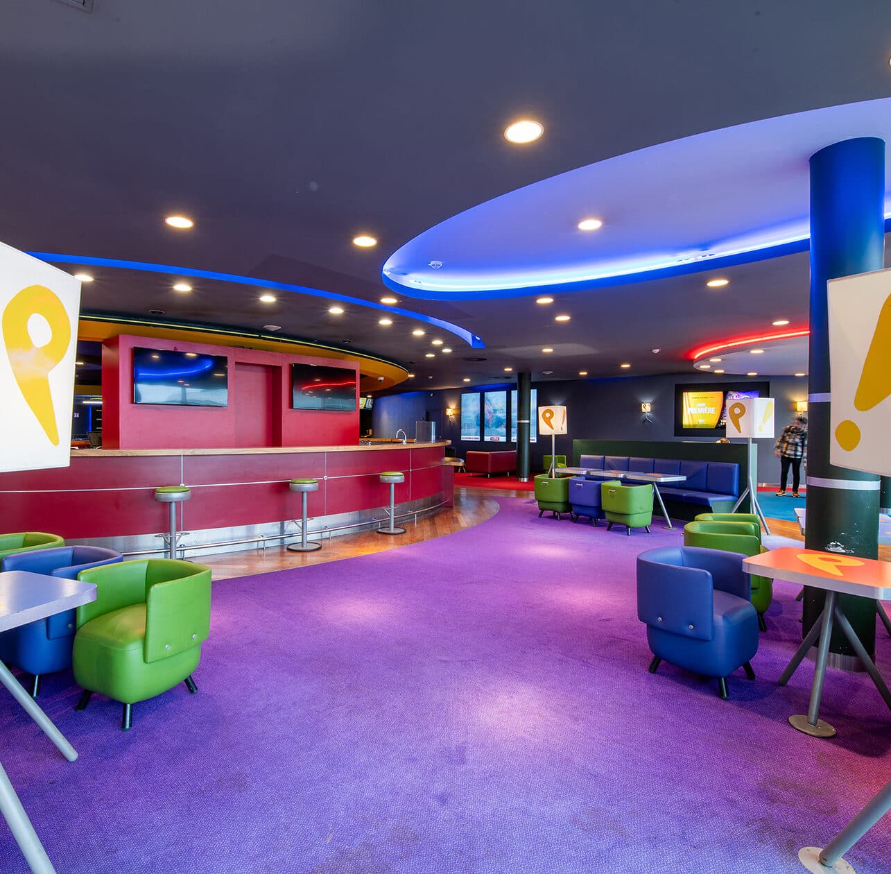 Espace cocktail du cinéma, éclairé par des néons colorés et aménagé avec du mobilier de réception vert et violet. Le bar est en fond.
