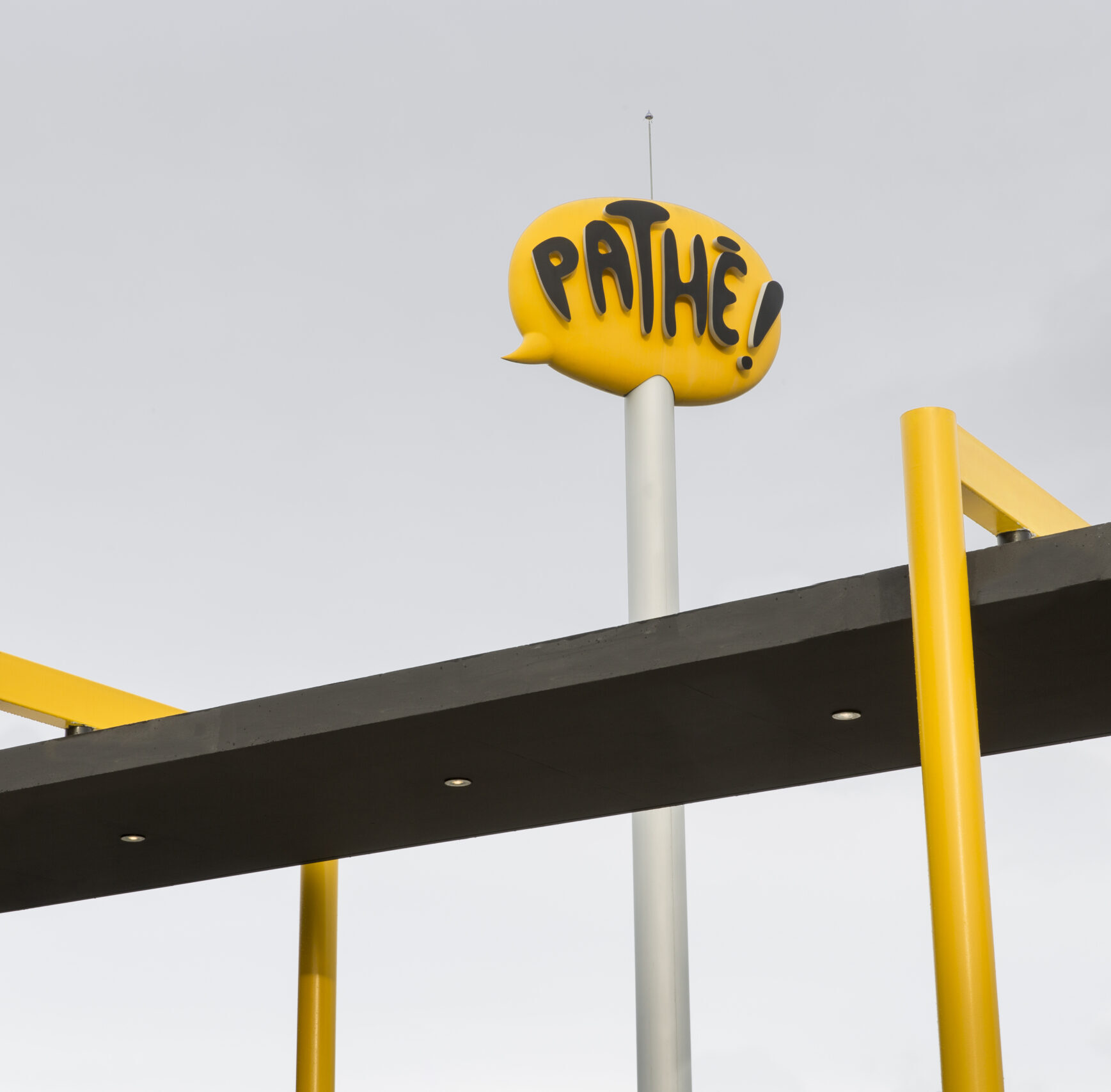 Enseigne jaune en forme de bulle de dialogue avec le mot “Pathe!” en lettres noires, installée en hauteur sur la façade moderne d’un bâtiment, sous un ciel gris clair