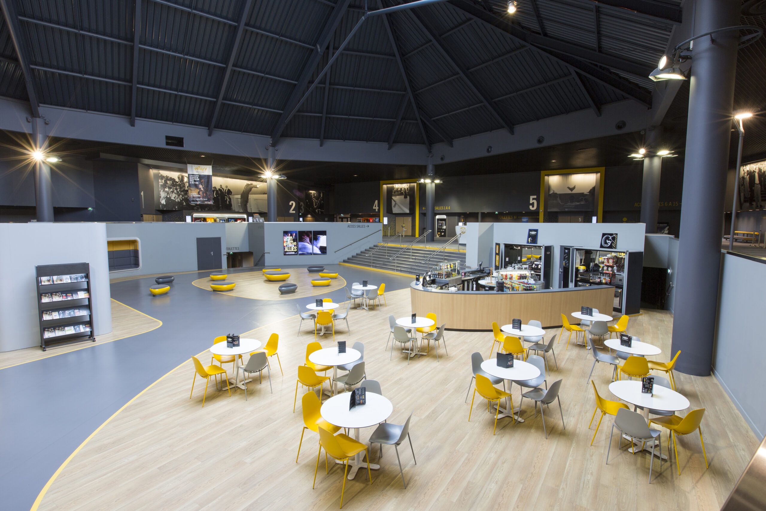Hall central d'un cinéma avec des tables entourées de chaises jaunes et grises, un comptoir de restauration, des escaliers menant aux salles, et un plafond haut en structure métallique.