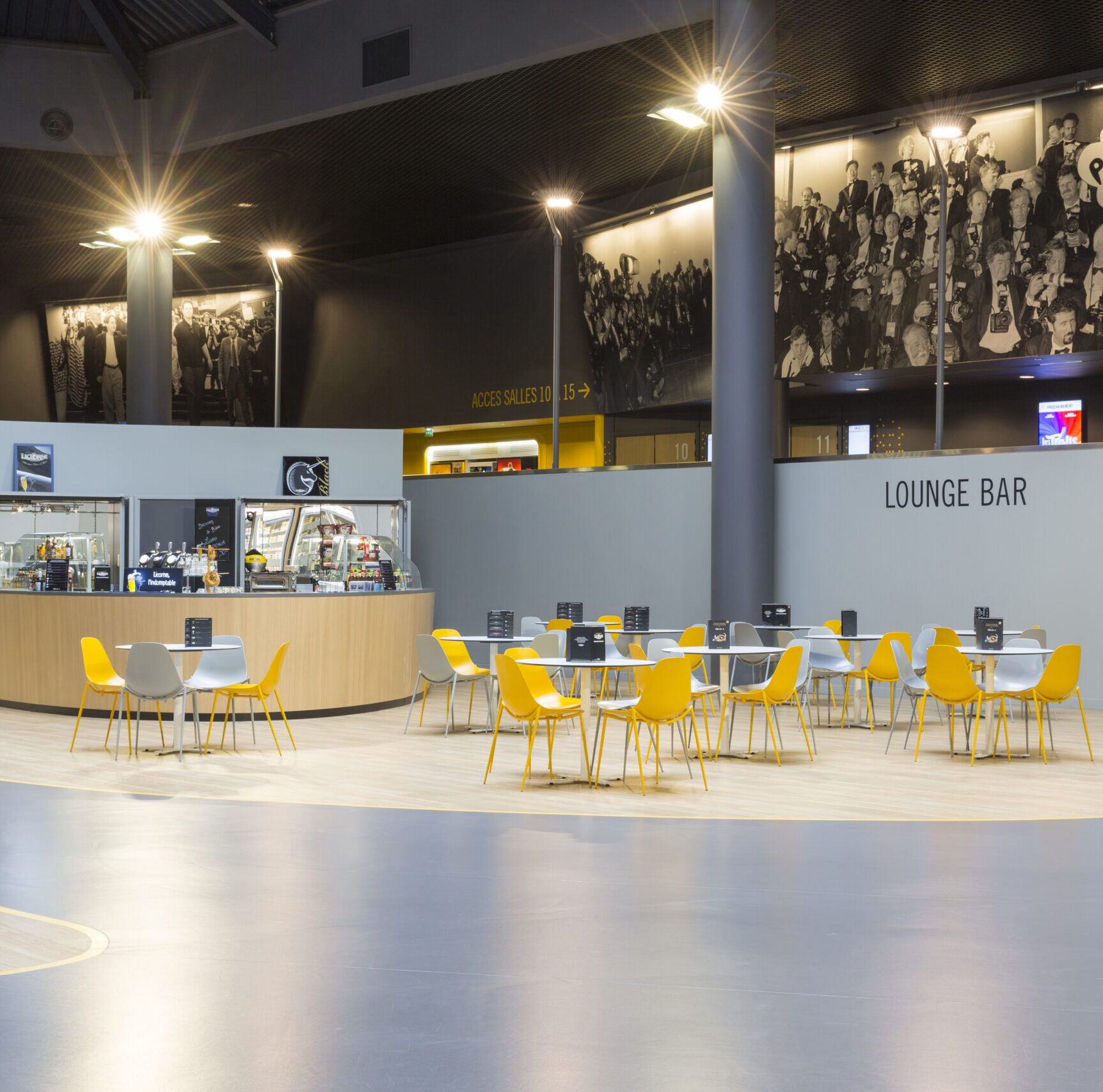 Hall central d'un cinéma avec des tables entourées de chaises jaunes et grises, et un comptoir de restauration.