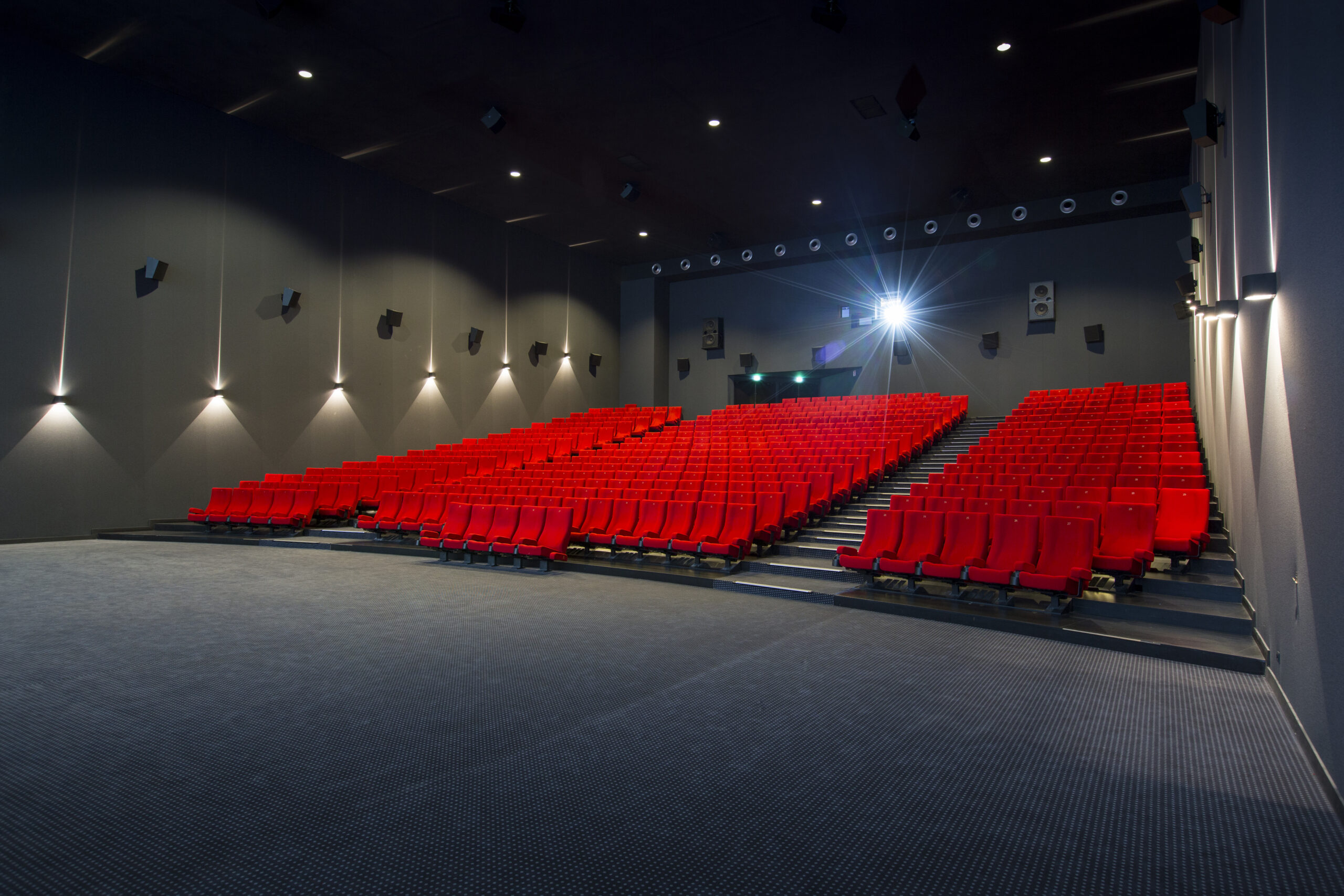 Vue latérale de la salle de cinéma montrant les fauteuils en velours rouges et l’agencement des rangées ainsi qu'une partie de son espace scénique.
