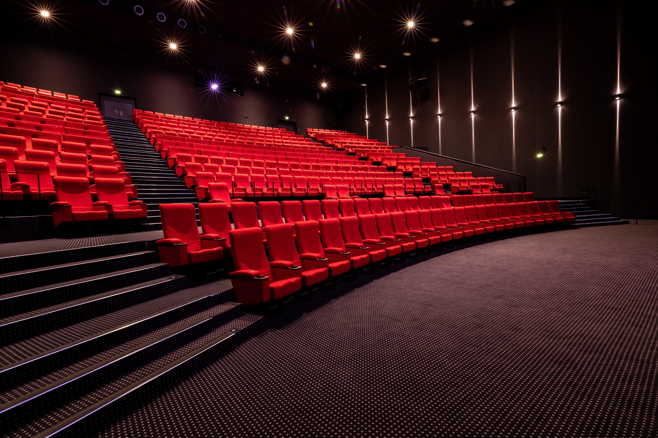 Vue latérale de la salle de cinéma montrant les fauteuils en velours rouges et l’agencement des rangées ainsi qu'une partie de son espace scénique.