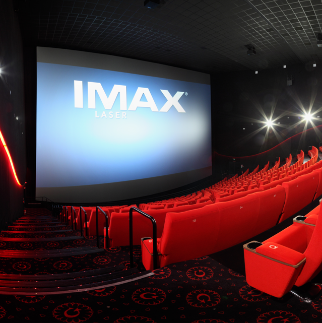 Vue de profil d'une salle de cinéma équipée de la technologie IMAX montrant les fauteuils en velours rouges et l’agencement des rangées ainsi que son espace scénique.