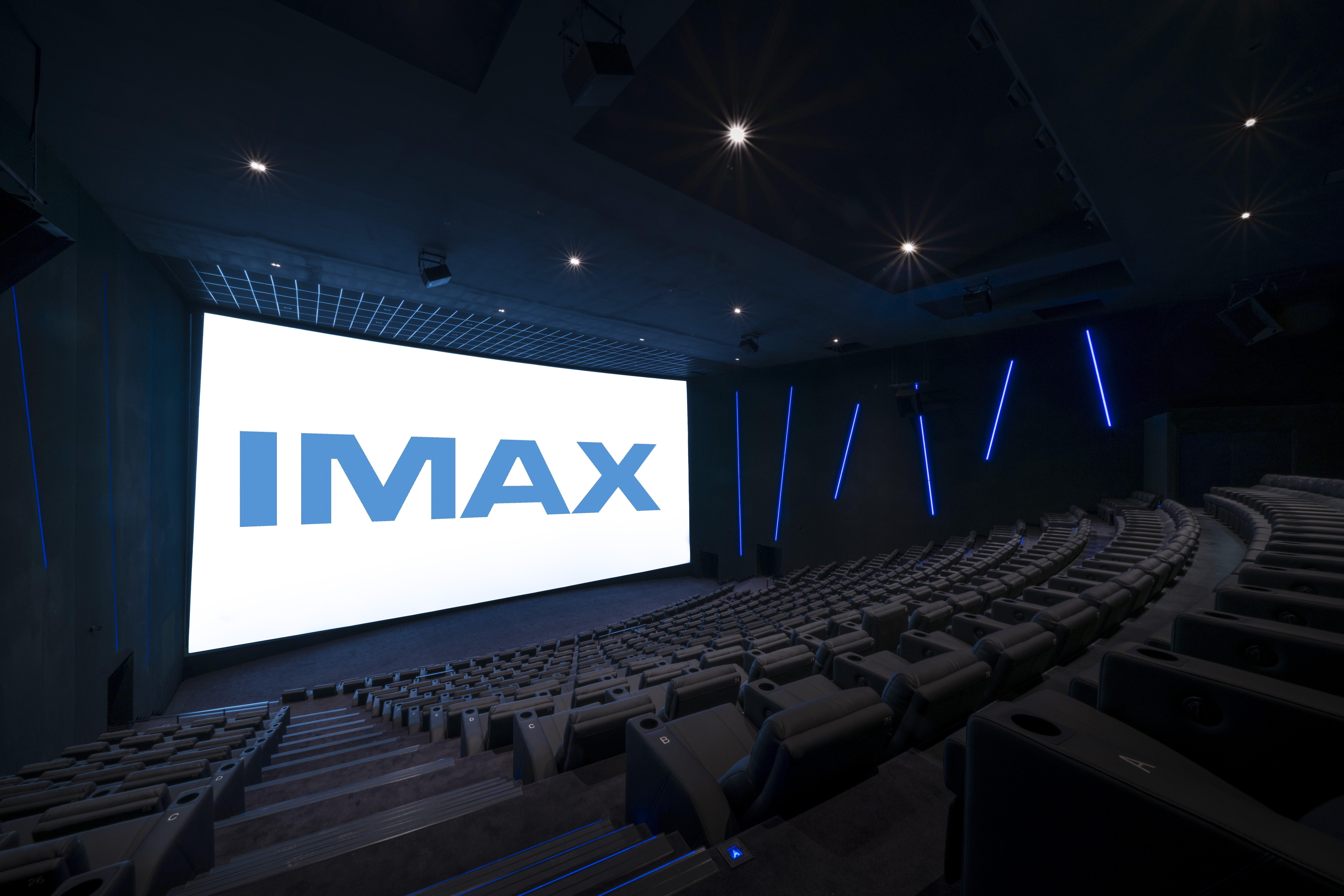 Vue de derrière d'une salle de cinéma équipée de la technologie IMAX montrant les fauteuils en cuirs noirs et l’agencement des rangées ainsi que son espace scénique.
