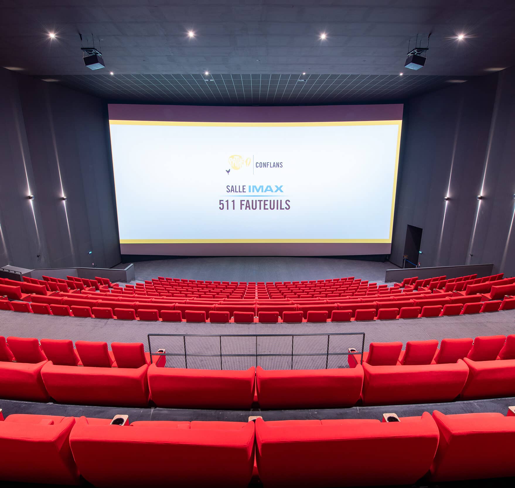Vue depuis l’arrière de la salle IMAX du cinéma Pathé Conflans avec rangées de fauteuils en velours rouges orientés vers l’écran.