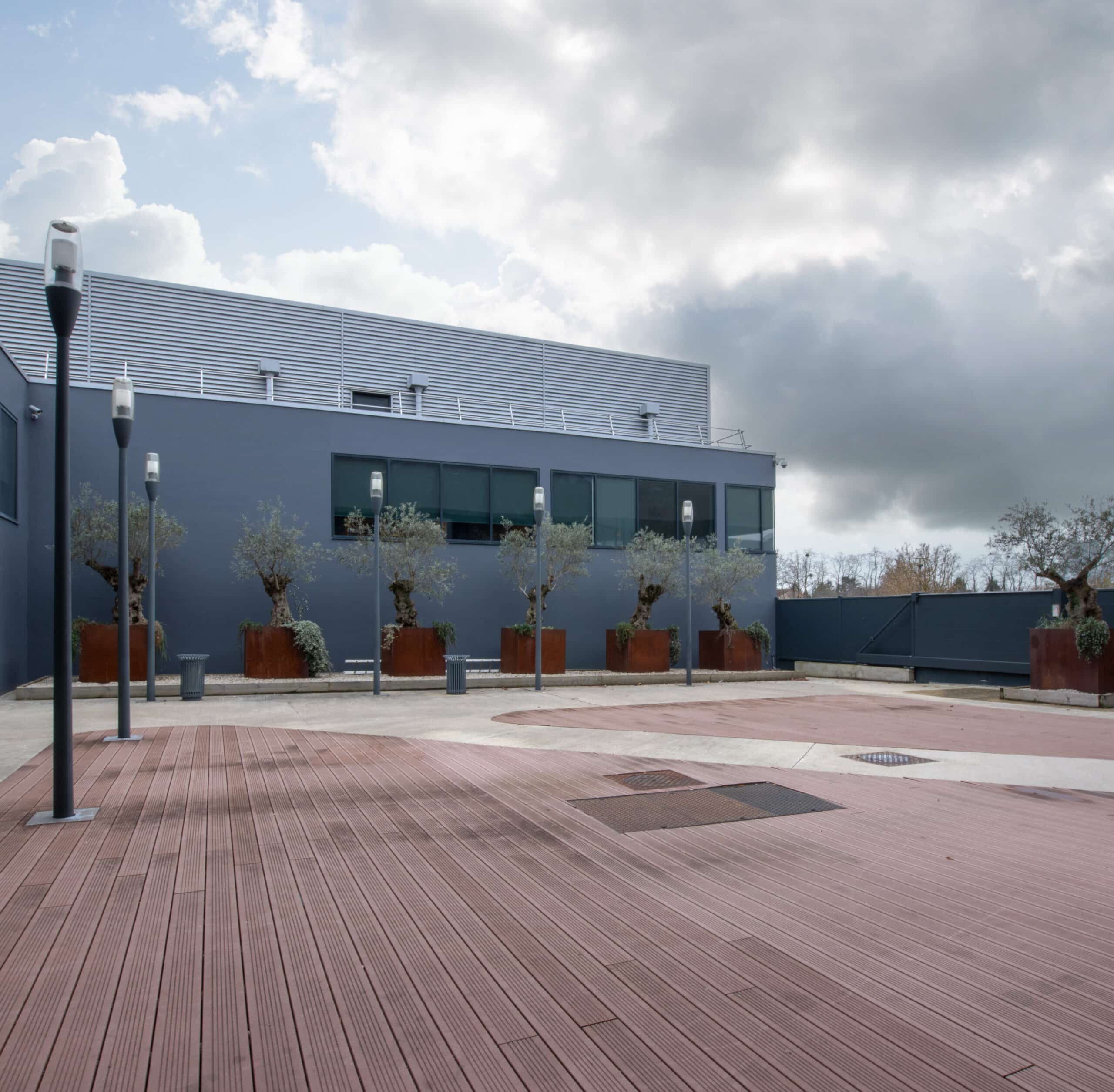 Cour extérieure avec un sol en bois et béton, entourée de lampadaires modernes et de jardinières en métal rouillé contenant des oliviers. La façade grise d’un bâtiment aux larges fenêtres est visible en arrière-plan, sous un ciel nuageux.