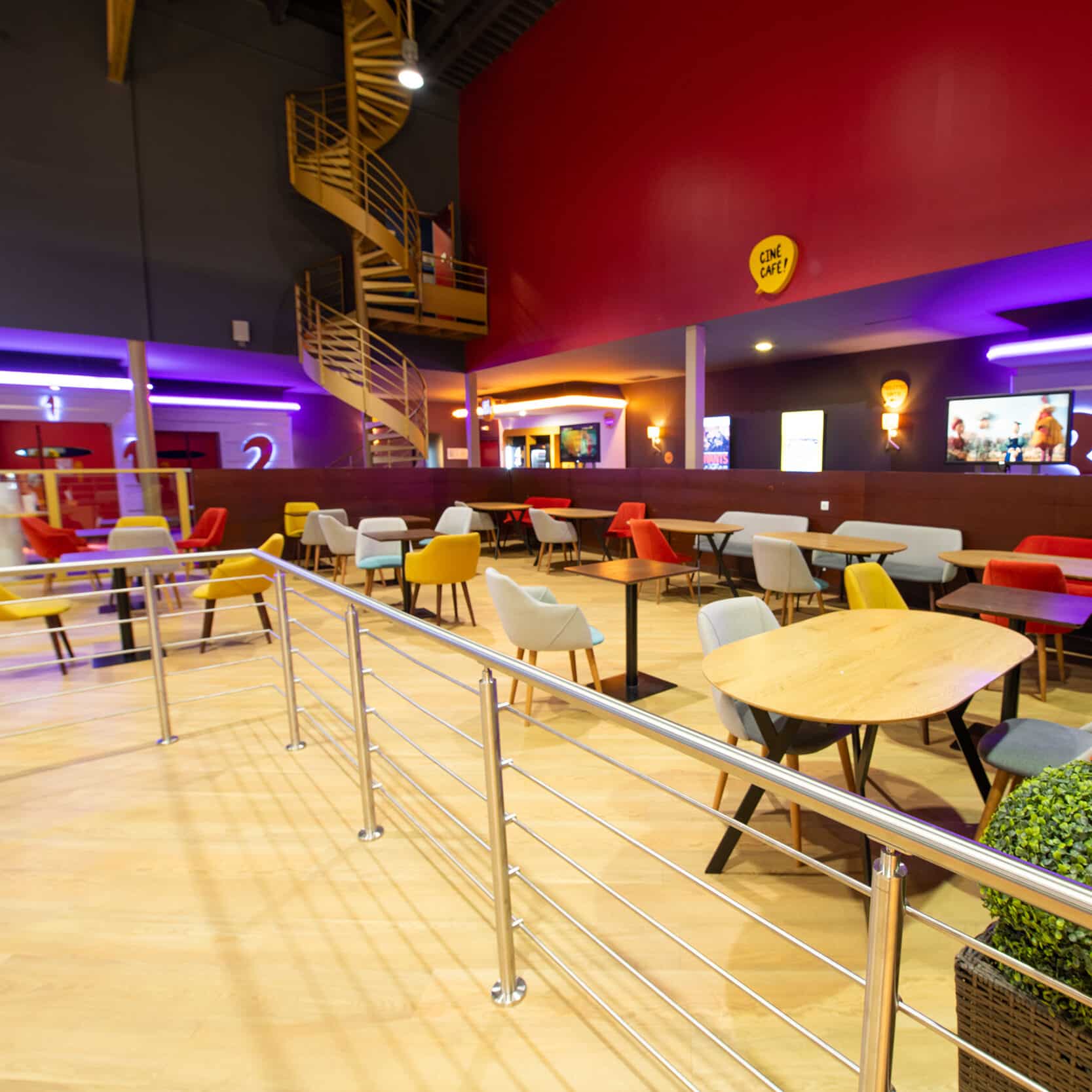 Espace café dans un cinéma avec des chaises colorées, des tables en bois, un éclairage lumineux et un escalier en colimaçon en arrière-plan.