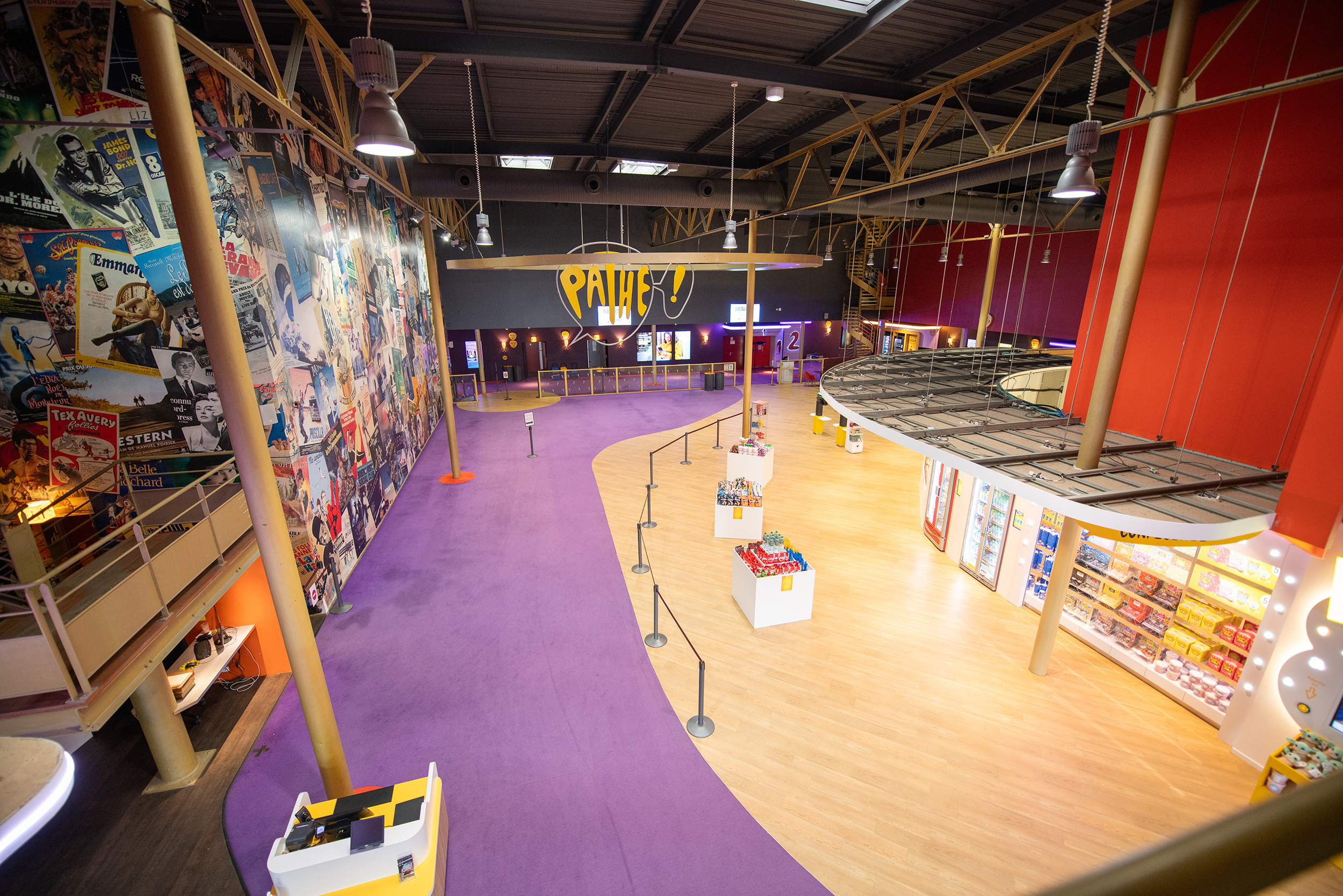 Hall spacieux et coloré dans un cinéma, avec un tapis violet, des étagères de confiseries, un mur décoré d'affiches de films, et des zones d'accès aux salles.