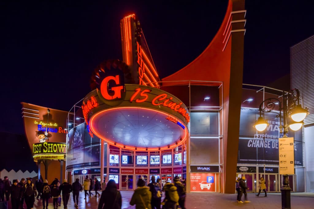 Façade lumineuse d'un cinéma avec enseigne circulaire éclairée en rouge, affiches de films, et architecture moderne à l'entrée, entourée de visiteurs le soir.