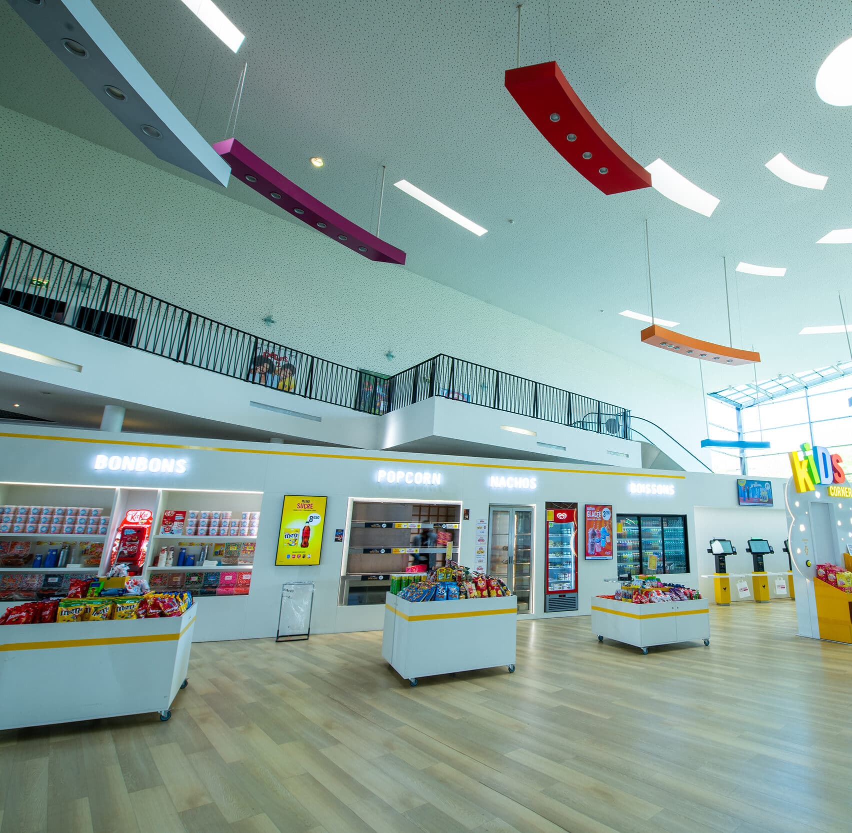 Espace confiserie lumineux dans un cinéma avec des étagères de bonbons, popcorns et boissons, des couleurs vives suspendues au plafond, et un sol en bois clair.