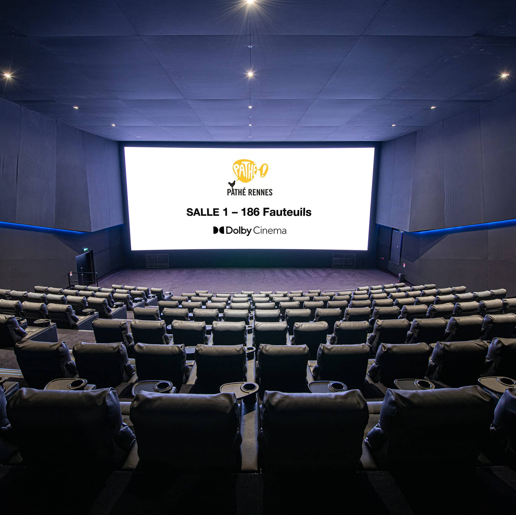 Salle 1 du cinéma de Rennes, sombre et moderne avec rangées de sièges en cuir, éclairage discret sur les murs et projecteur allumé au fond, créant une ambiance immersive.