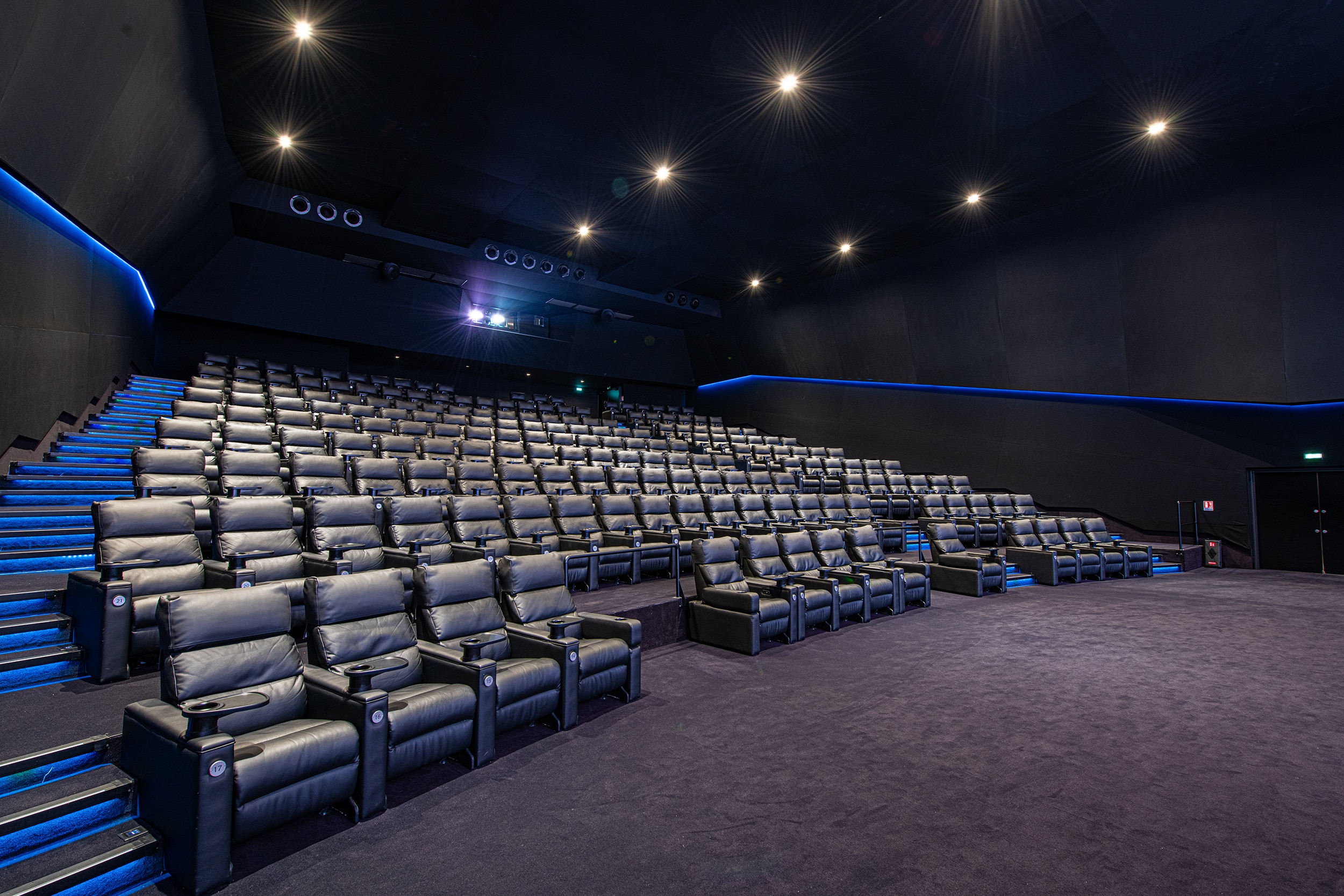 Salle de cinéma sombre et moderne avec rangées de sièges en cuir, éclairage sur les murs et projecteur allumé au fond, créant une ambiance immersive.