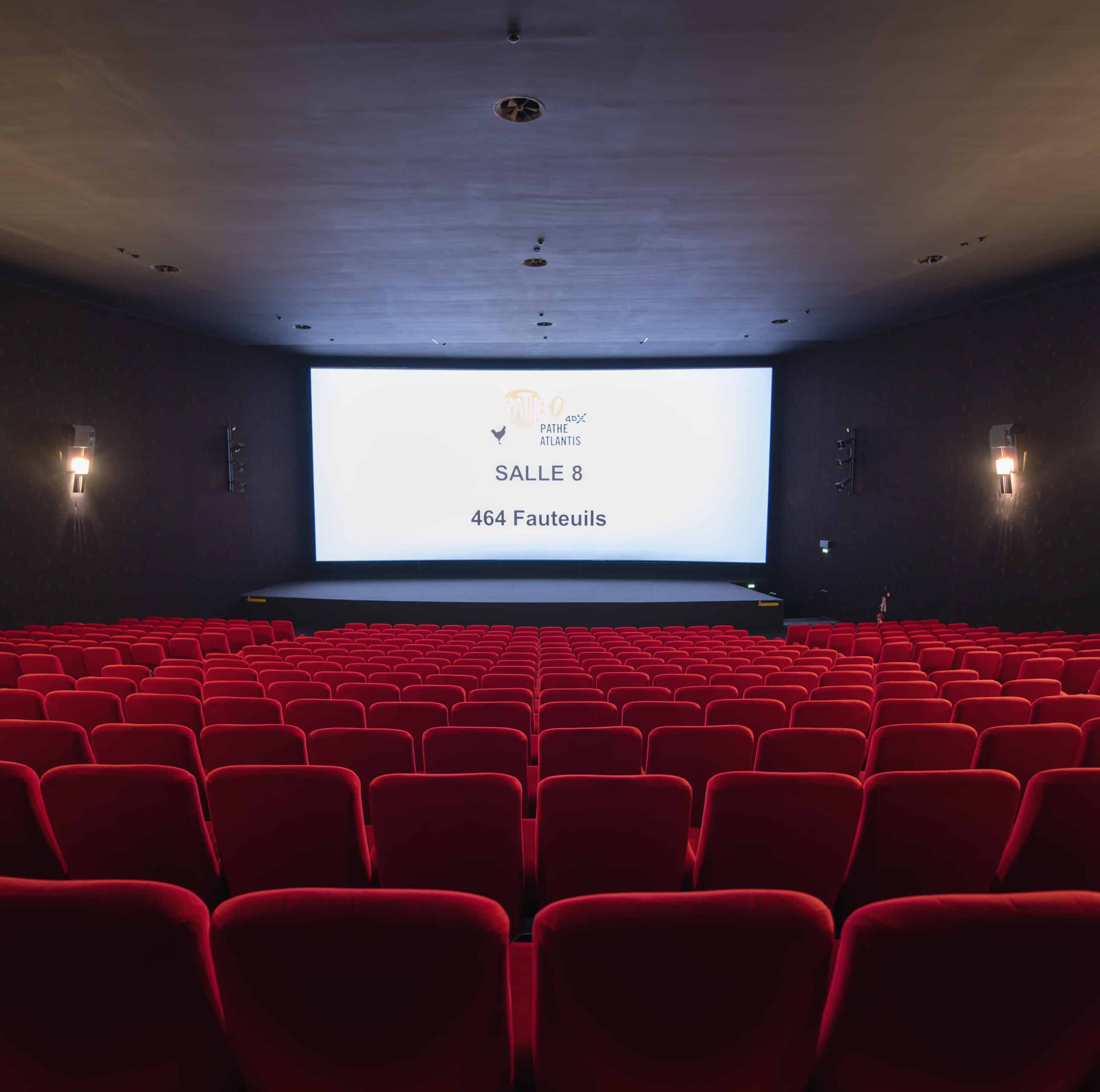 Vue depuis l’arrière de la salle 8 du cinéma Pathé Atlantis avec rangées de fauteuils en velours rouges orientés vers l’écran.