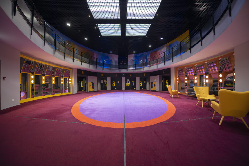 Hall circulaire coloré avec tapis violet et orange, numéros de salles visibles, fauteuils jaunes et éclairage moderne.