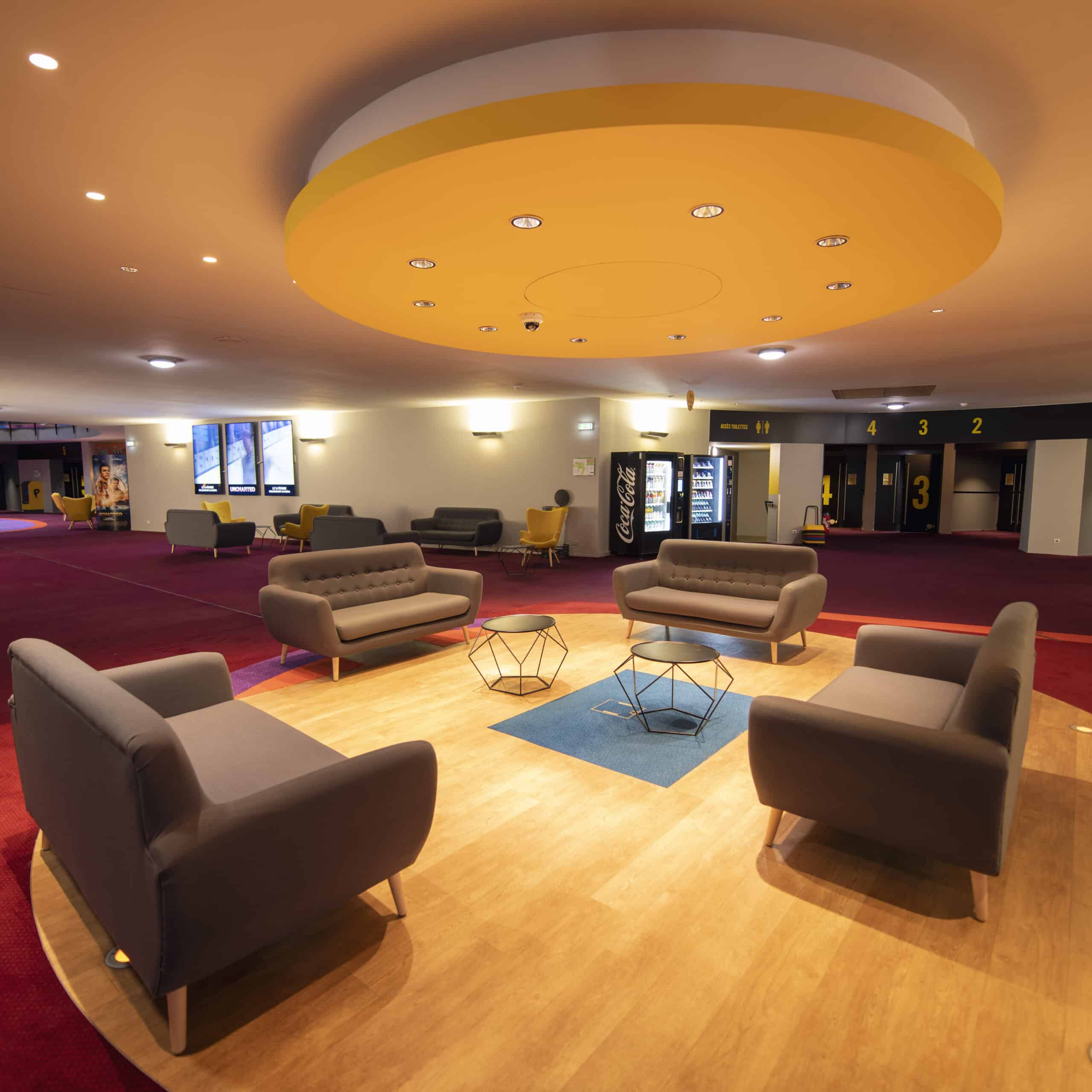 Espace de réception confortable du cinéma avec fauteuils gris, tables modernes et plafonnier jaune.