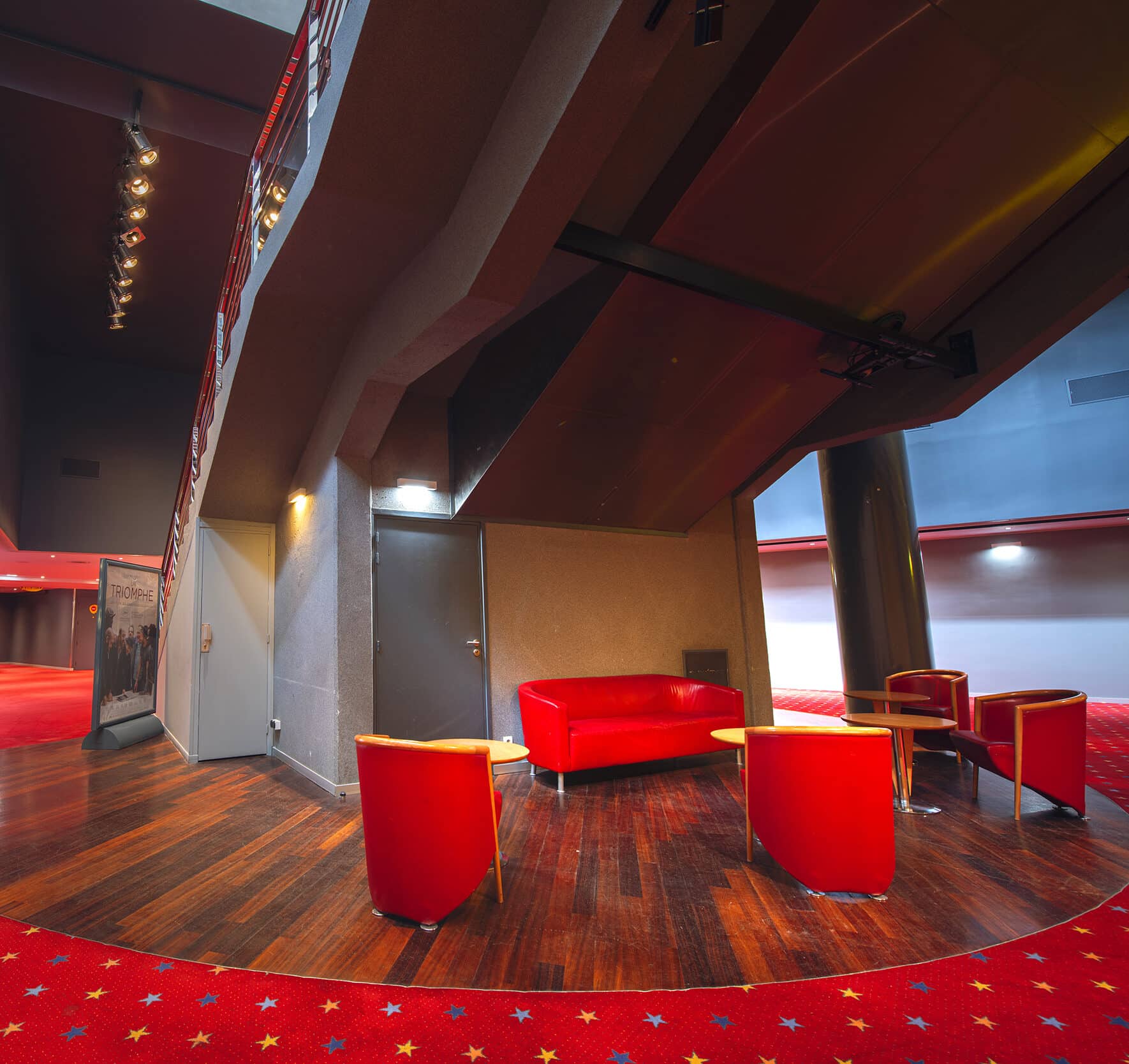 Espace de réception dans un cinéma avec des fauteuils et un canapé rouges, un sol en bois, et un escalier moderne.