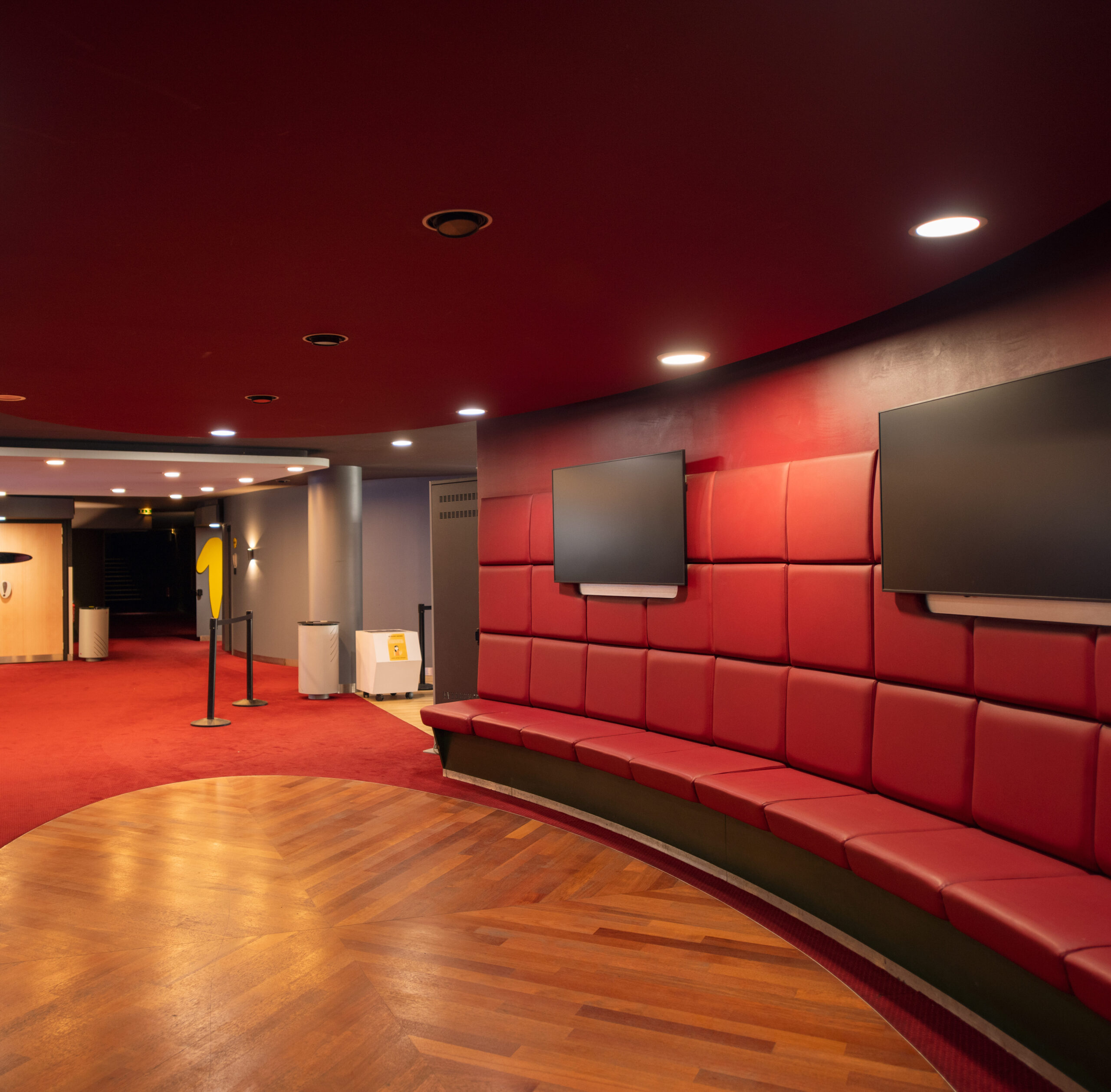 Espace de réception situé dans le hall du cinéma, éclairé par une lumière tamisée et aménagé avec des canapés dans les tons rouges.