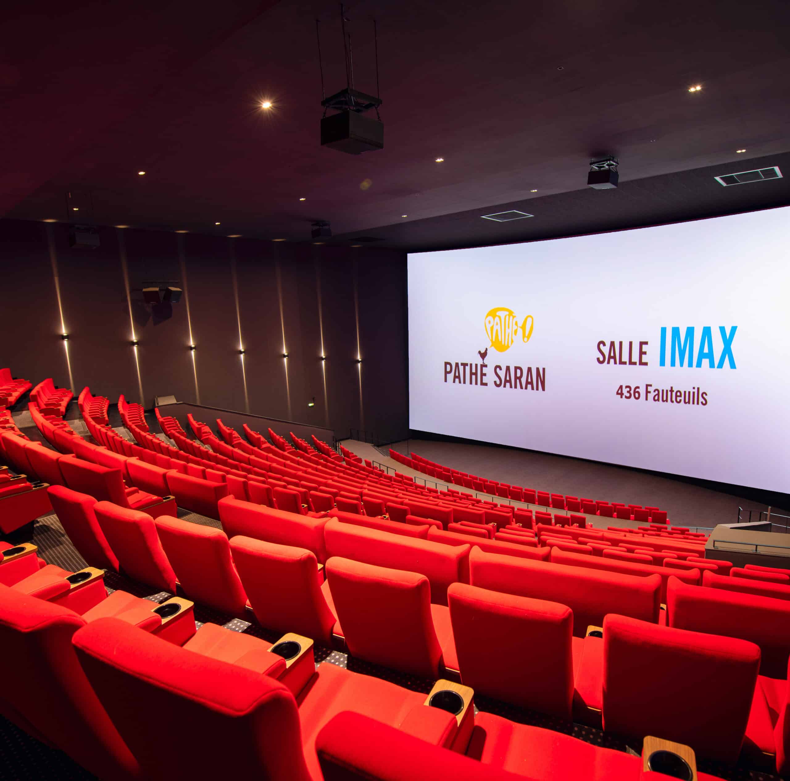 Vue depuis l’arrière d'une salle de cinéma équipée de la technologie IMAX avec rangées de fauteuils en velours rouges orientés vers l’écran.